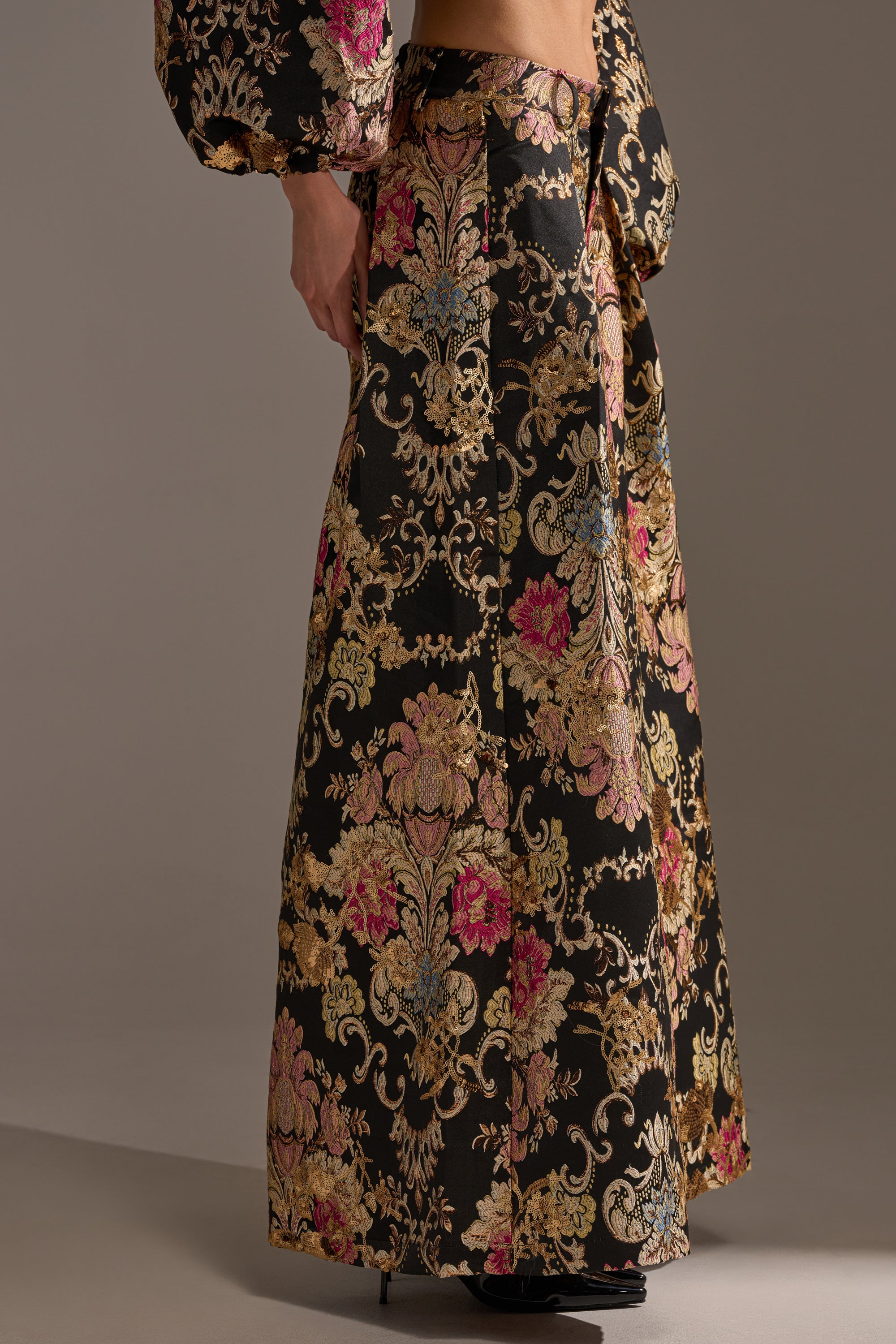 A person wears the LUXE ENERGY PALAZZO PANT in black, gold, pink, and blue with an ornate brocade print. Matching puffed sleeves and black heeled shoes complete this elegant look.