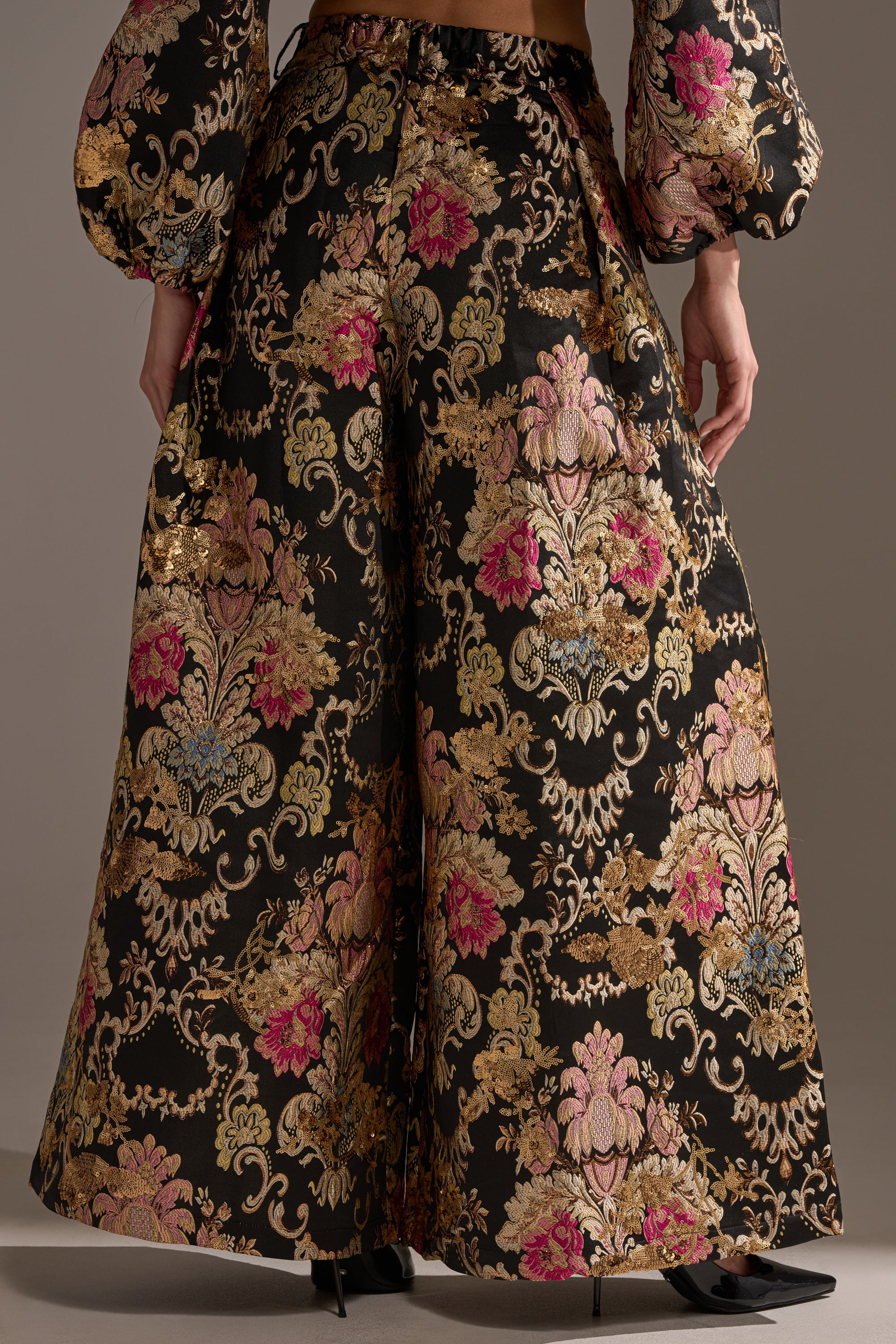 A person wears the LUXE ENERGY PALAZZO PANT, featuring a brocade print in gold, pink, and metallic hues on black. Styled with a matching long-sleeve puffy top and black pointed-toe heels for a chic look.