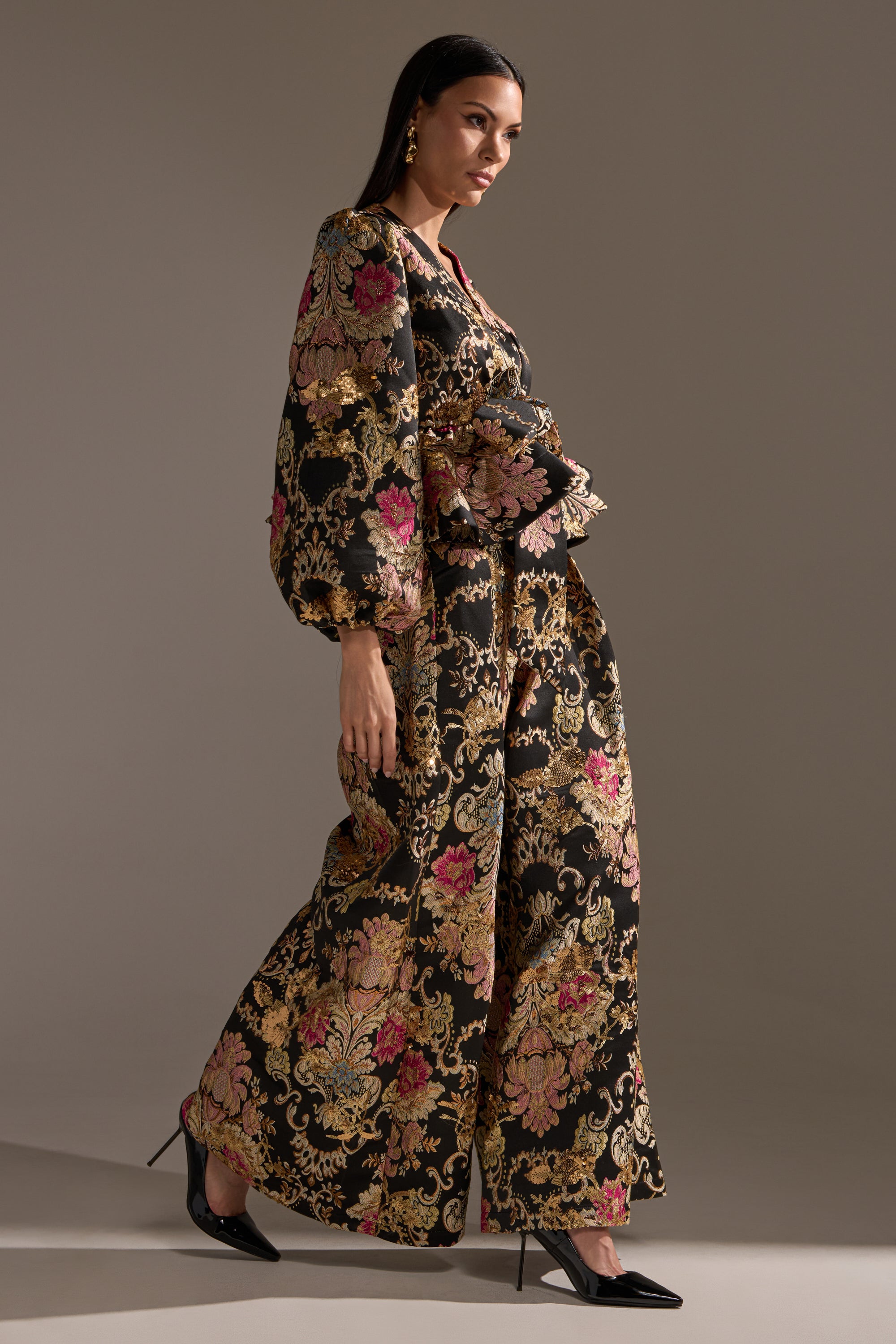 A woman in the LUXE ENERGY PALAZZO PANT suit, featuring brocade-print pants and dramatic sleeves, strides confidently in black pointed-toe heels against a neutral background.