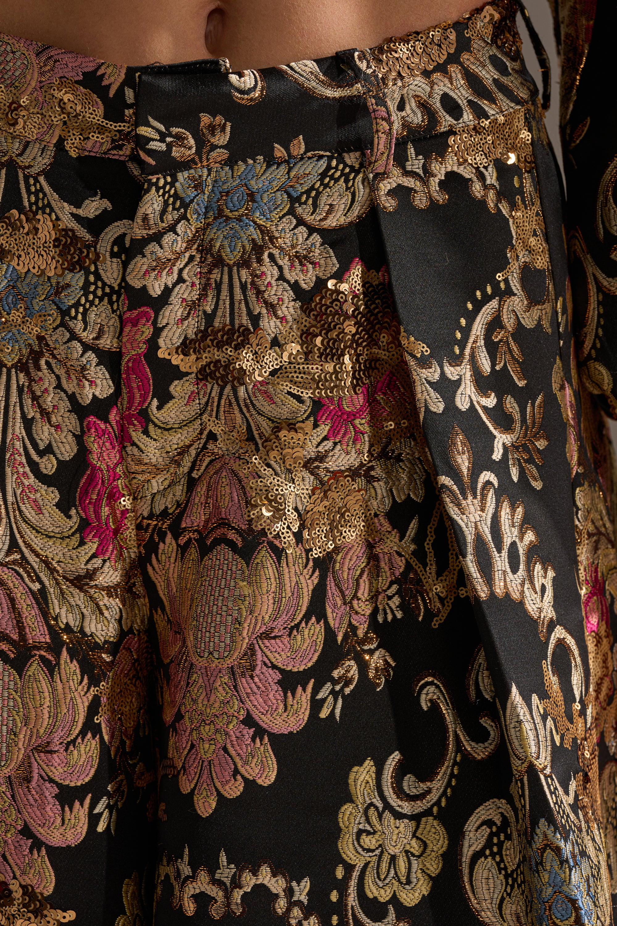 Close-up of the LUXE ENERGY PALAZZO PANT showcasing a detailed gold, pink, blue, and beige floral brocade on black, with pleats and a wide-leg silhouette for added texture.