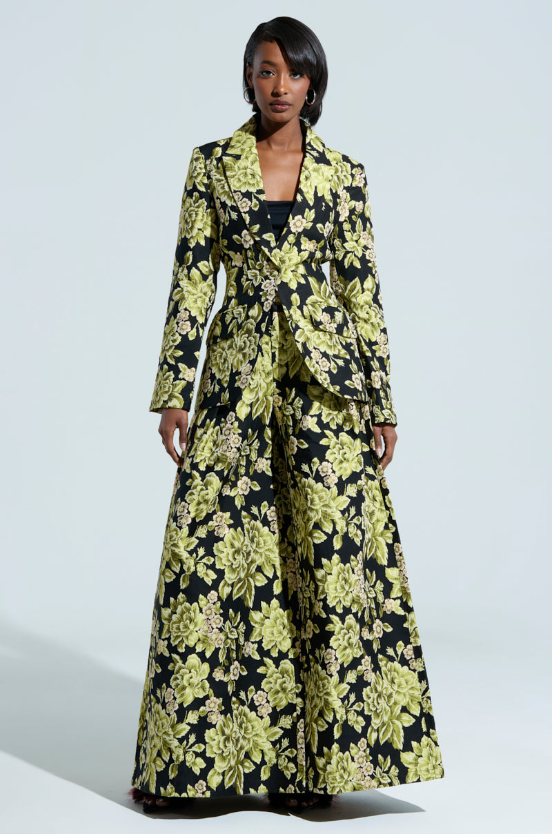 A woman dons a tailored black suit adorned with the elaborate BUY MYSELF FLOWERS BROCADE TROUSER, showcasing a vibrant yellow floral brocade pattern. The ensemble includes a structured blazer paired with wide-legged trousers. Set against a plain, light background, the bold design of her outfit captures the essence of being able to buy herself flowers every day.