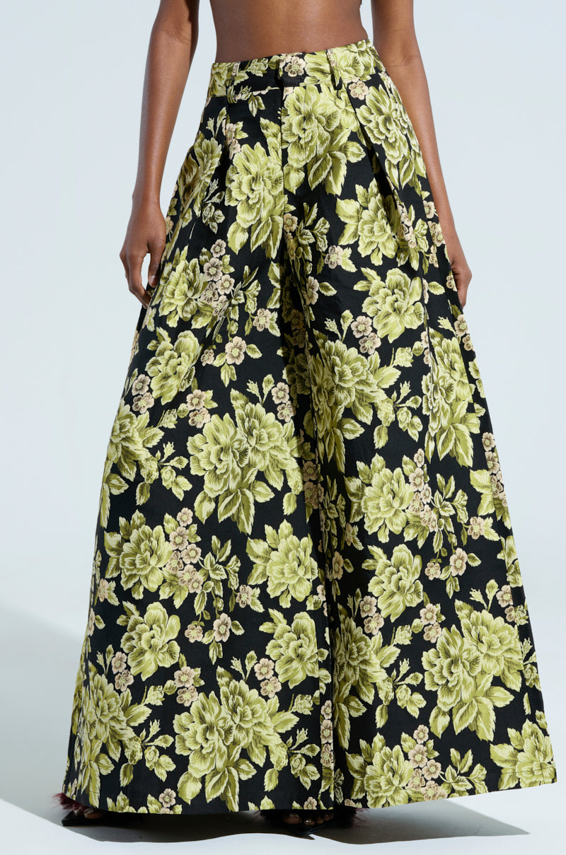 Someone is showcasing the "BUY MYSELF FLOWERS BROCADE TROUSER," which features wide-legged brocade trousers adorned with a striking floral pattern in green and yellow hues on a dark backdrop. The emphasis is on the pants’ high waist and pleated design.