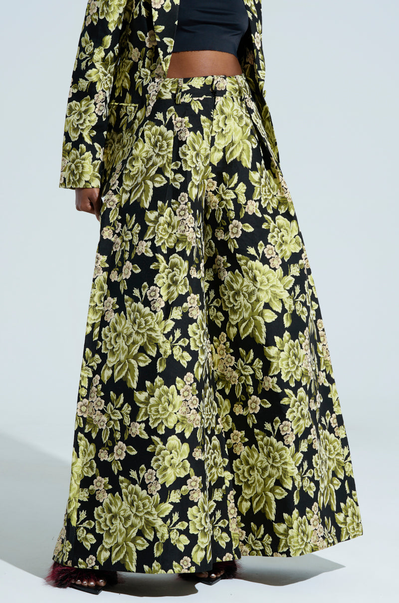 A person is wearing the BUY MYSELF FLOWERS BROCADE TROUSER paired with a matching jacket, both featuring an intricate flower pattern in bold green and black brocade. The photo focuses on the lower half of the ensemble against a plain backdrop.