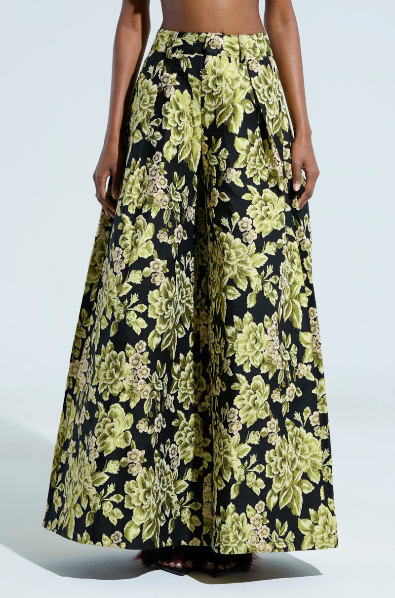 A person wearing the BUY MYSELF FLOWERS BROCADE TROUSER, which features wide-legged, high-waisted brocade with a bold floral pattern in shades of yellow and green on a black background. The pleats create a voluminous silhouette. The upper body is not visible.