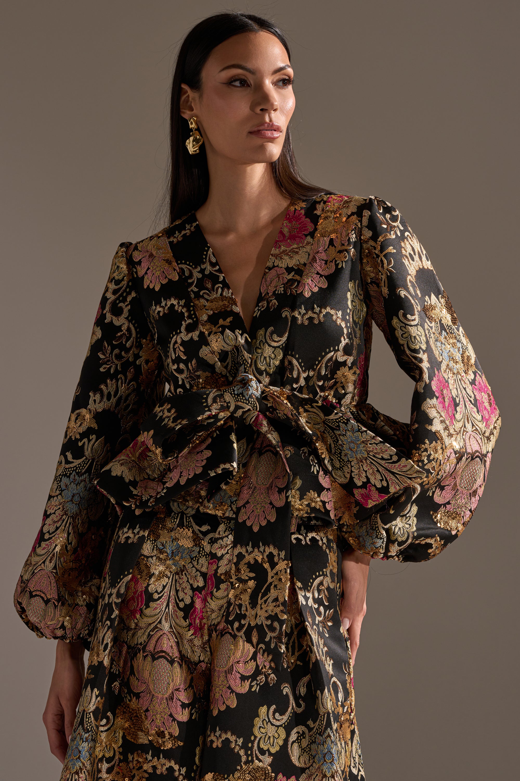 Wearing the LUXE ENERGY BROCADE BLOUSE, a woman poses against a neutral background. The black blouse is adorned with ornate gold, pink, and blue floral patterns and subtle metallic detailing. She looks to the side with one hand on her hip.