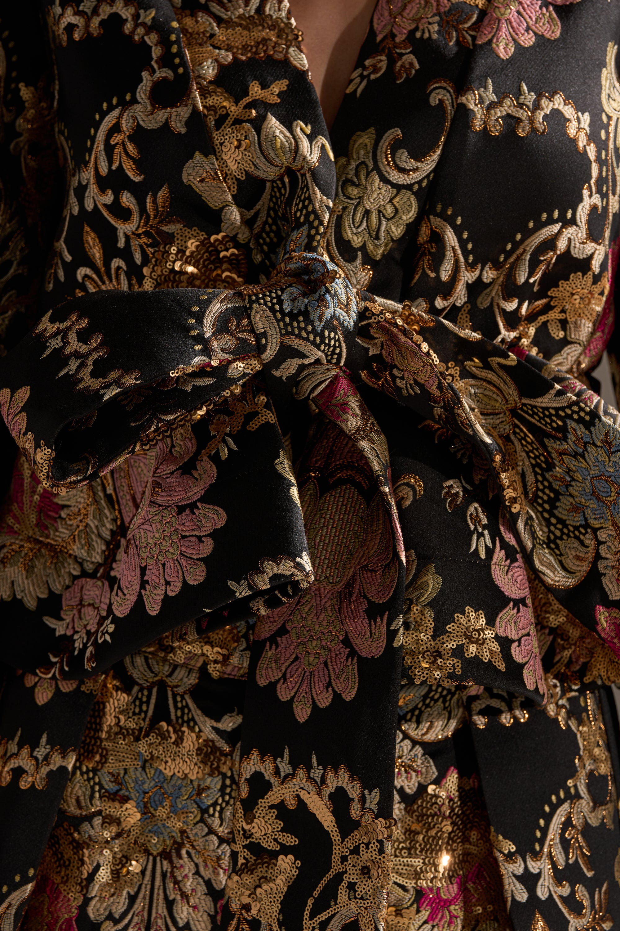 Close-up of the LUXE ENERGY BROCADE BLOUSE in black, featuring gold metallic details and pink-blue floral embroidery. A large bow at the center highlights its luxurious texture and intricate patterns.