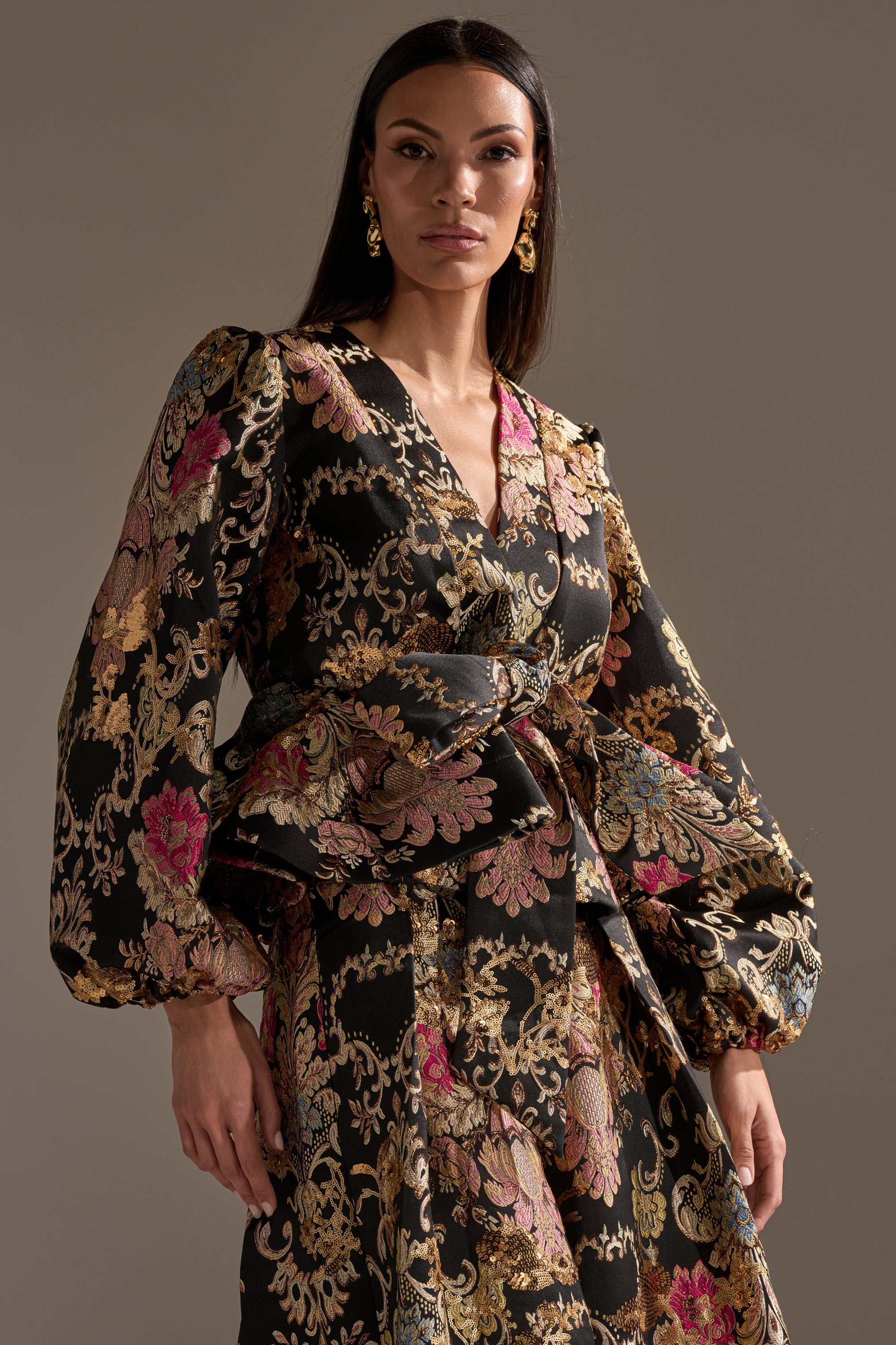A woman stands against a neutral background wearing the LUXE ENERGY BROCADE BLOUSE—black with gold metallic details, pink and beige floral patterns, puffed long sleeves, a cinched waist—paired with gold earrings and her straight brown hair down.