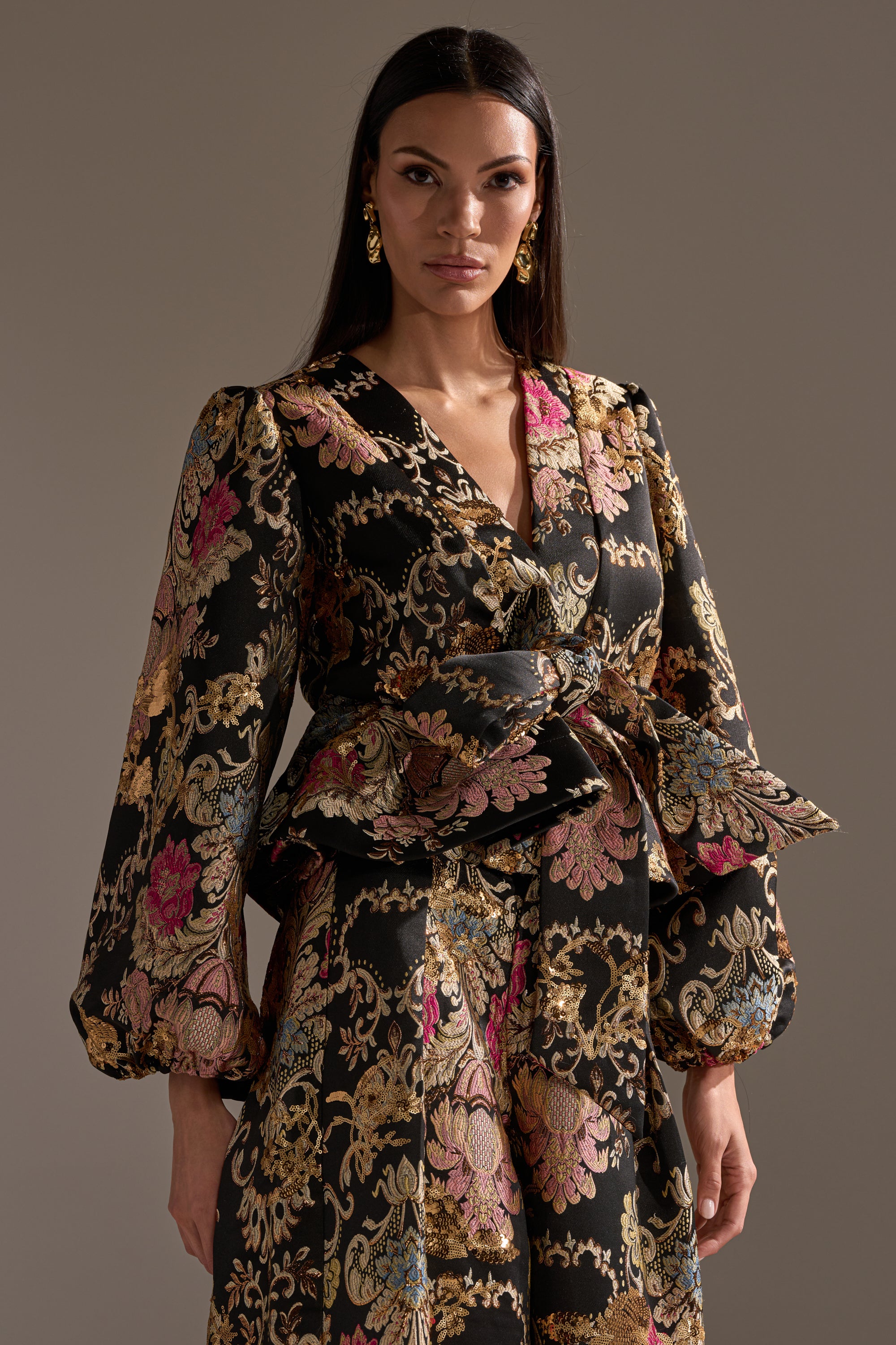 A woman with long dark hair wears the LUXE ENERGY BROCADE BLOUSE with gold metallic details, puffed long sleeves, a V-neckline, and a tied waist, standing against a plain neutral background.