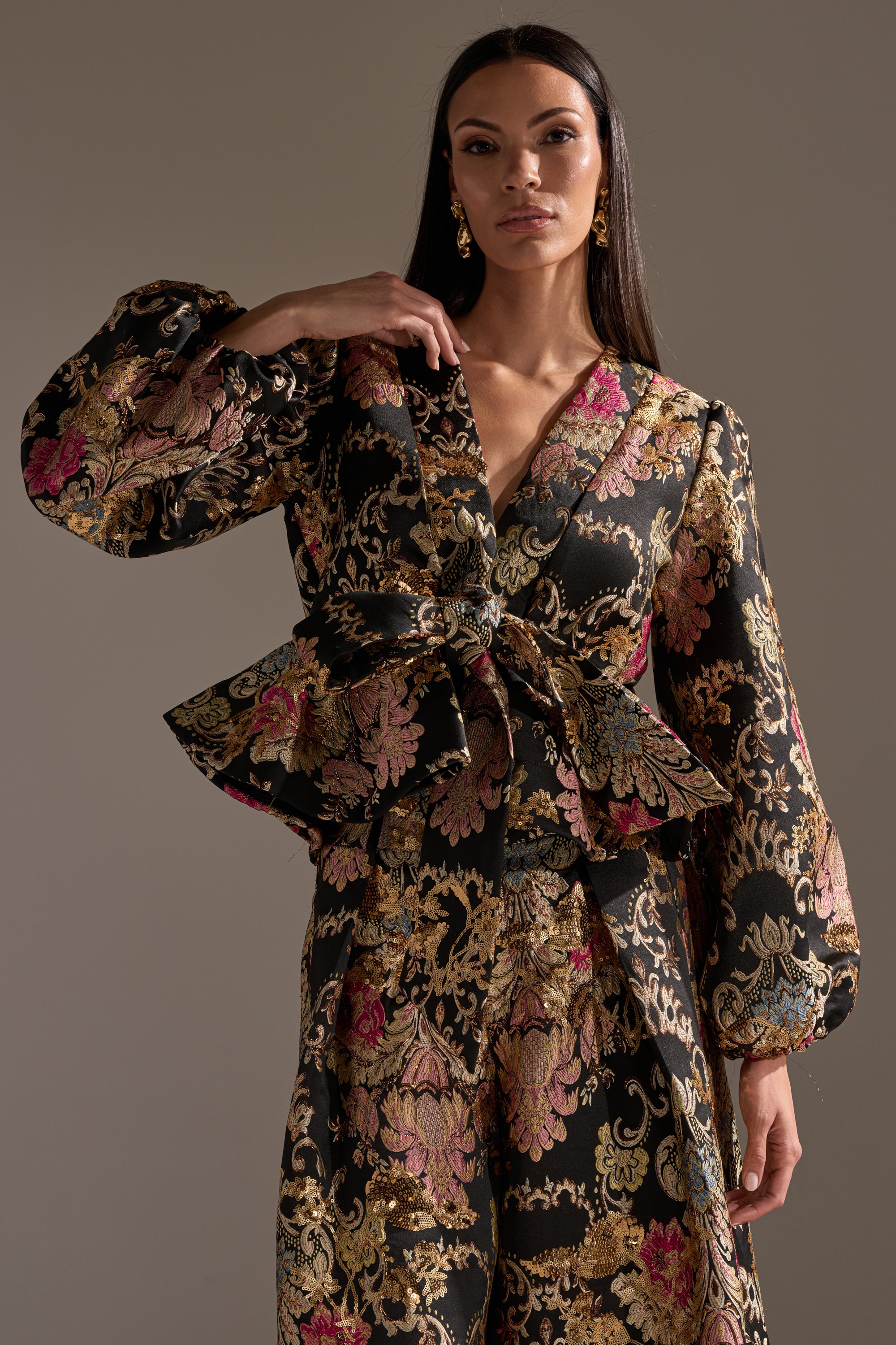 A woman models the LUXE ENERGY BROCADE BLOUSE, showcasing gold, pink, and beige floral patterns with voluminous sleeves and a statement waist bow. She has long dark hair and accessorizes with gold earrings.