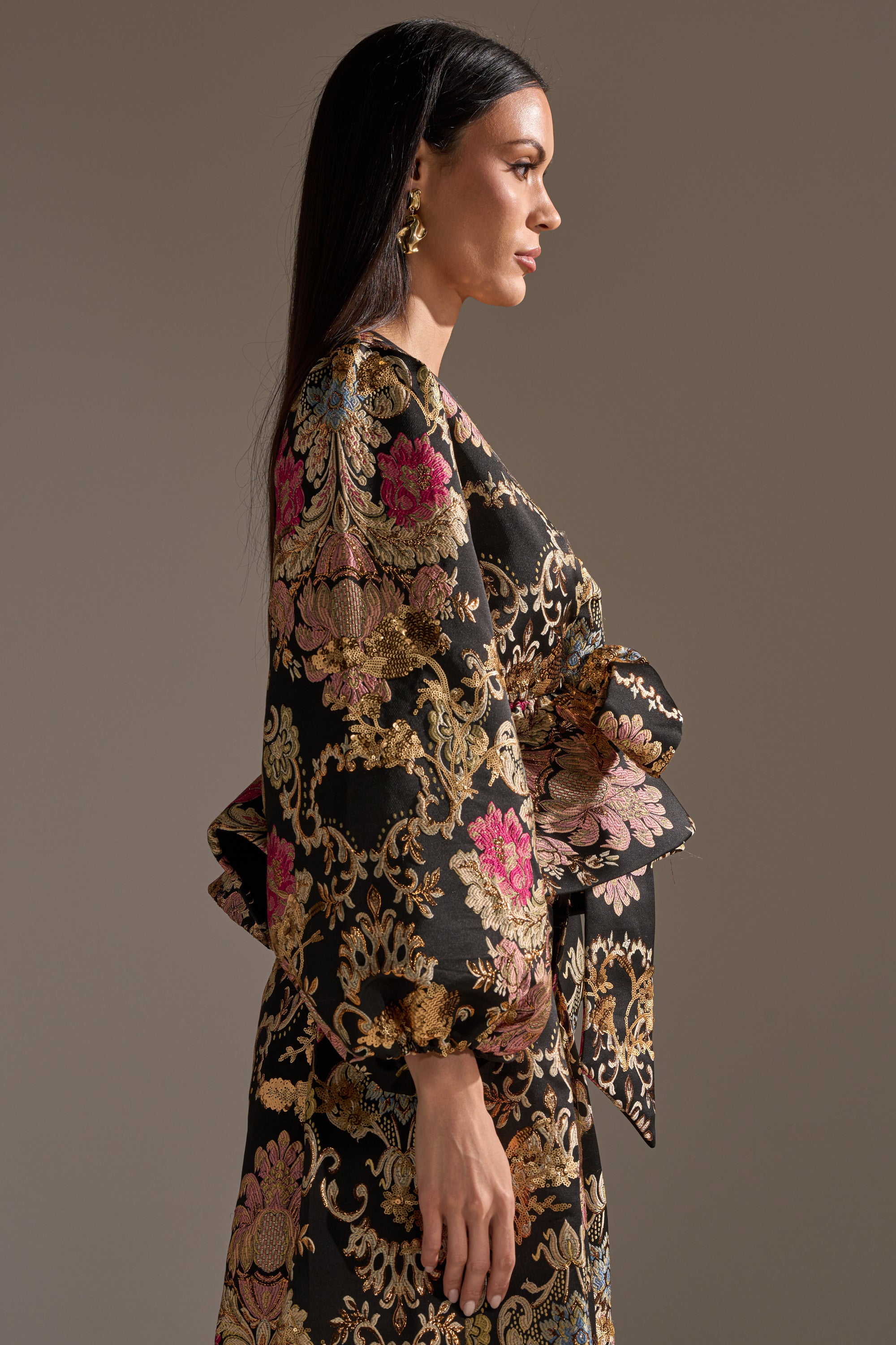 A woman with long dark hair stands in profile against a plain background, wearing the LUXE ENERGY BROCADE BLOUSE featuring gold, pink, and beige floral brocade and voluminous sleeves. She accessorizes with gold earrings.