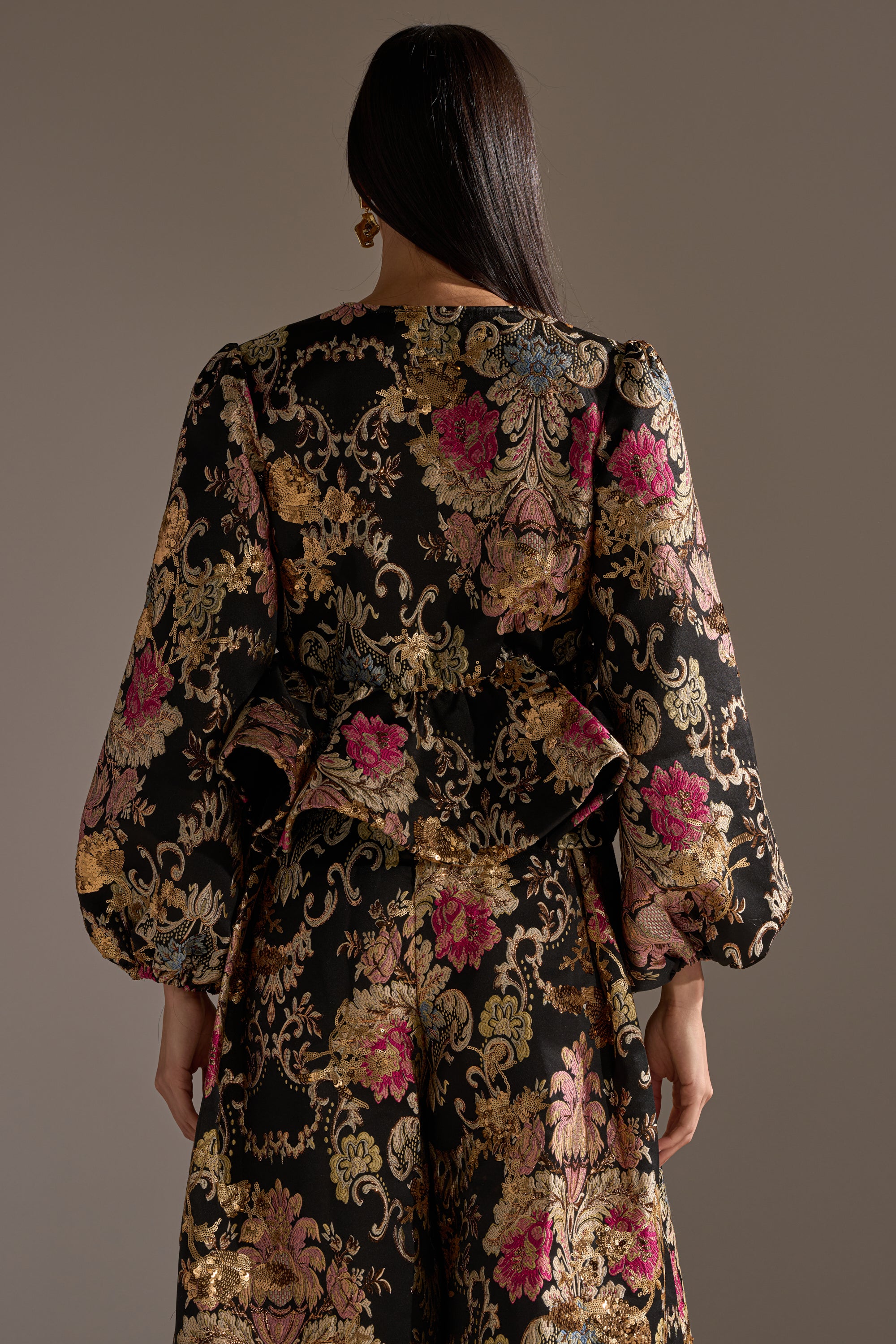 A person with long dark hair, seen from behind, wears the LUXE ENERGY BROCADE BLOUSE featuring gold, pink, and blue floral patterns with voluminous sleeves, standing against a plain brown background.