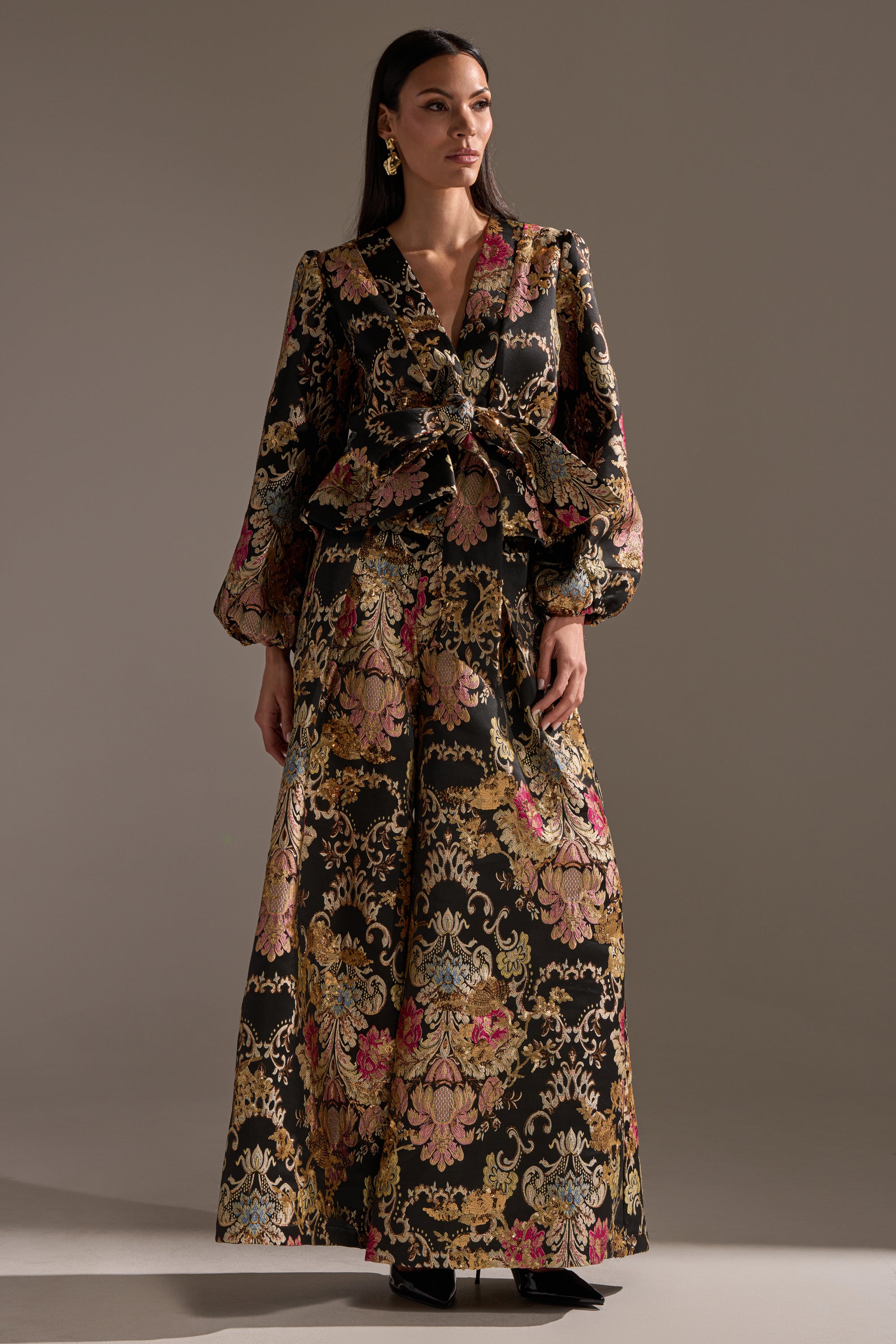 A woman stands in a studio wearing the Luxe Energy Brocade Blouse, featuring gold, pink, and green floral patterns, gold metallic detailing, a V-neck, balloon sleeves, peplum silhouette, large waist bow, and statement earrings.