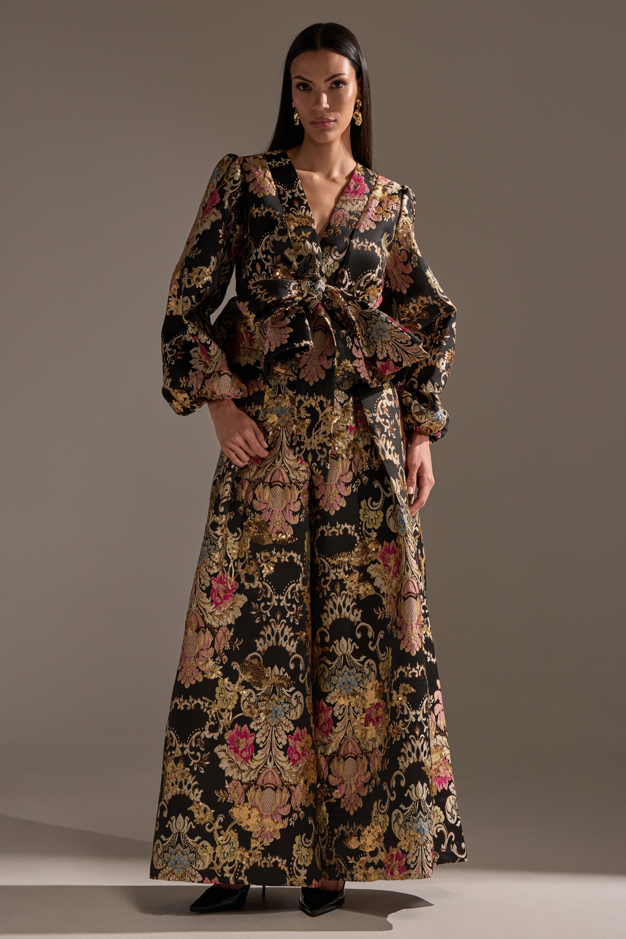 A woman stands in a studio wearing the LUXE ENERGY BROCADE BLOUSE, featuring gold metallic details, pink floral patterns, puffed sleeves, and a peplum silhouette. She pairs it with wide-leg trousers and black pointed shoes.