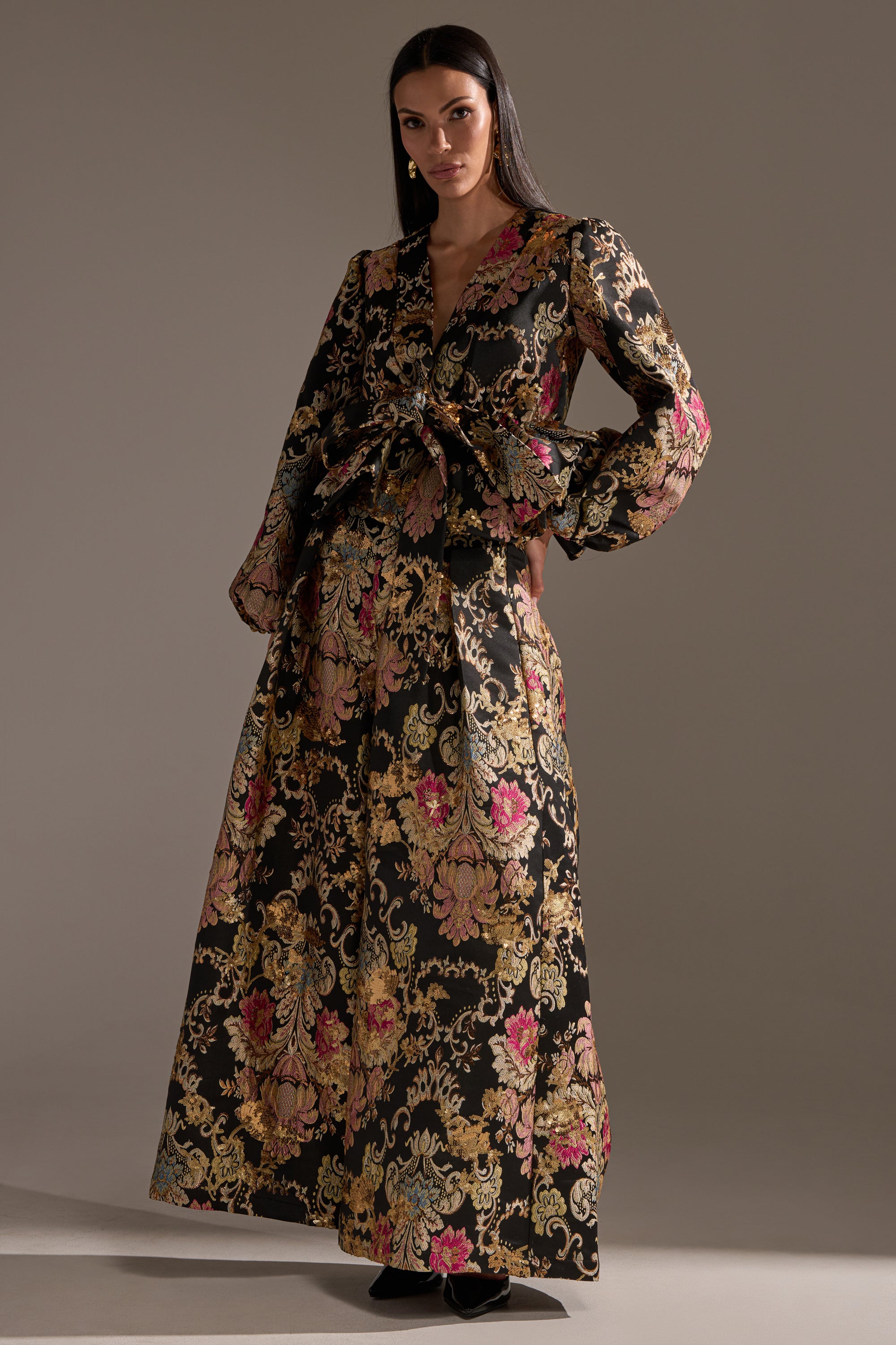 A woman wears the LUXE ENERGY BROCADE BLOUSE, featuring gold and pink floral brocade with gold metallic details, long sleeves, a deep V-neck, and a bow at the waist. She stands against a plain, softly lit background.
