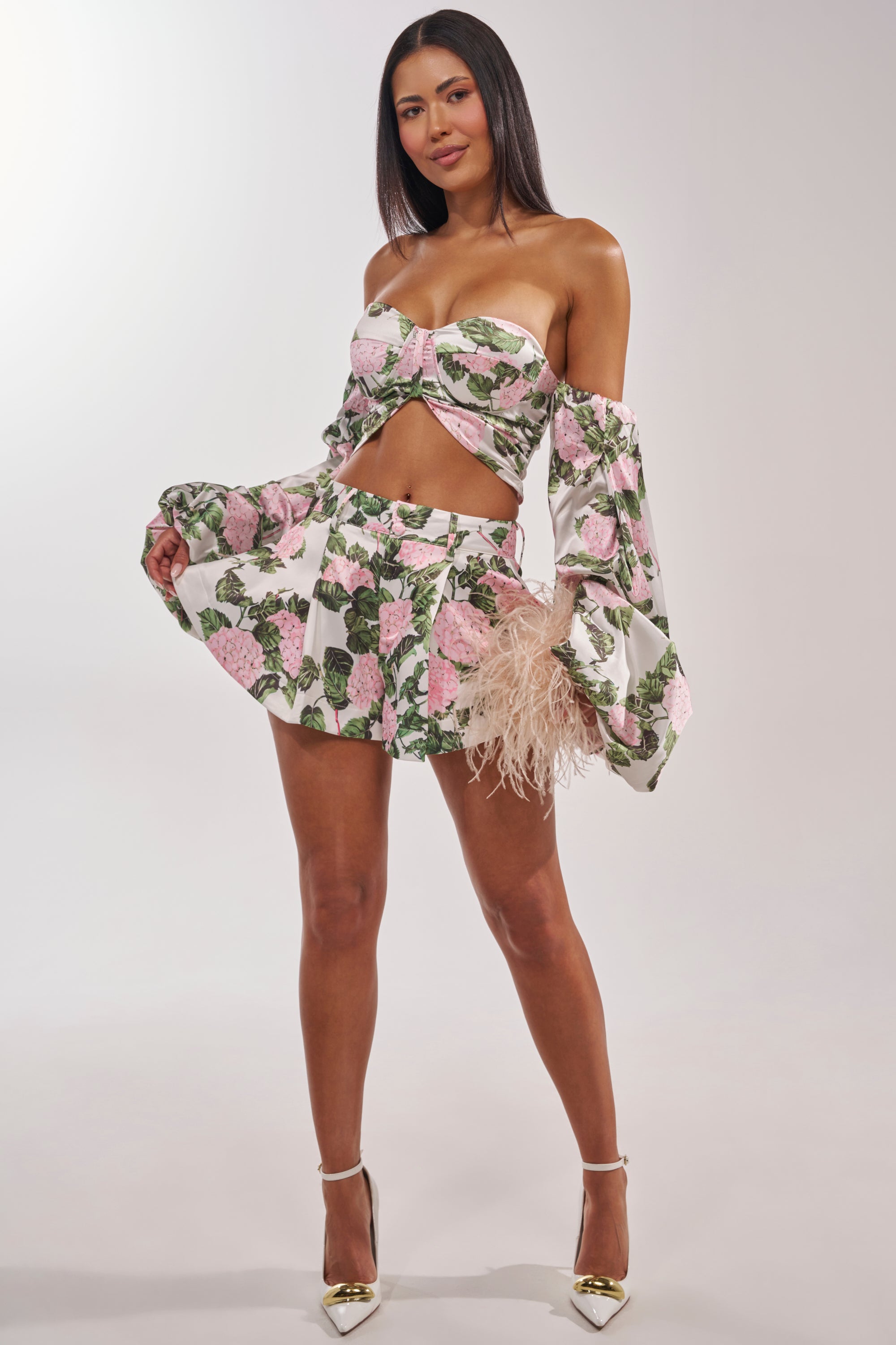 A woman stands confidently in the ONE OF A KIND FLORAL SHORT set—a matching floral crop top and flared mini shorts with pink and green patterns, puffed long sleeves, white high heels, straight long dark hair, and a neutral expression. 1
