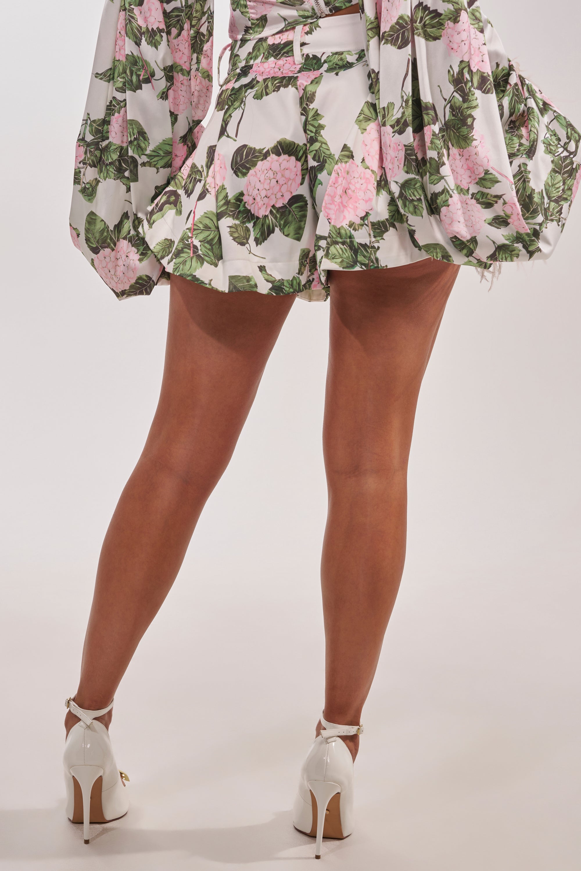 Person standing with legs crossed, wearing ONE OF A KIND FLORAL SHORT—satin shorts featuring pink and green patterns—paired with flowy sleeves and white high-heeled shoes against a light background. 5