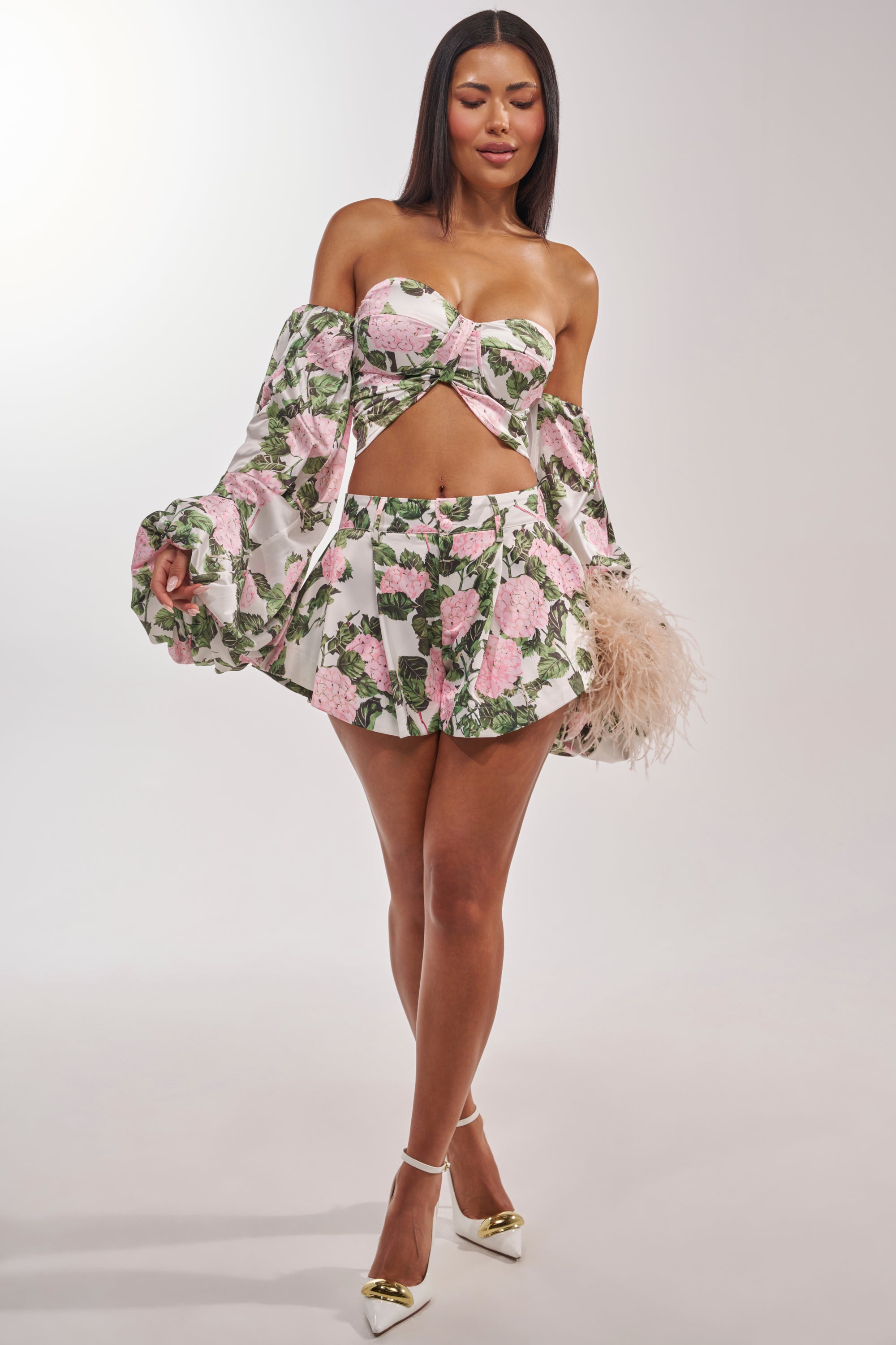 A woman models the ONE OF A KIND FLORAL SHORT, a two-piece set with a strapless crop top and flared mini shorts in pink and green floral print, styled with white heels and a feathered handbag. 6