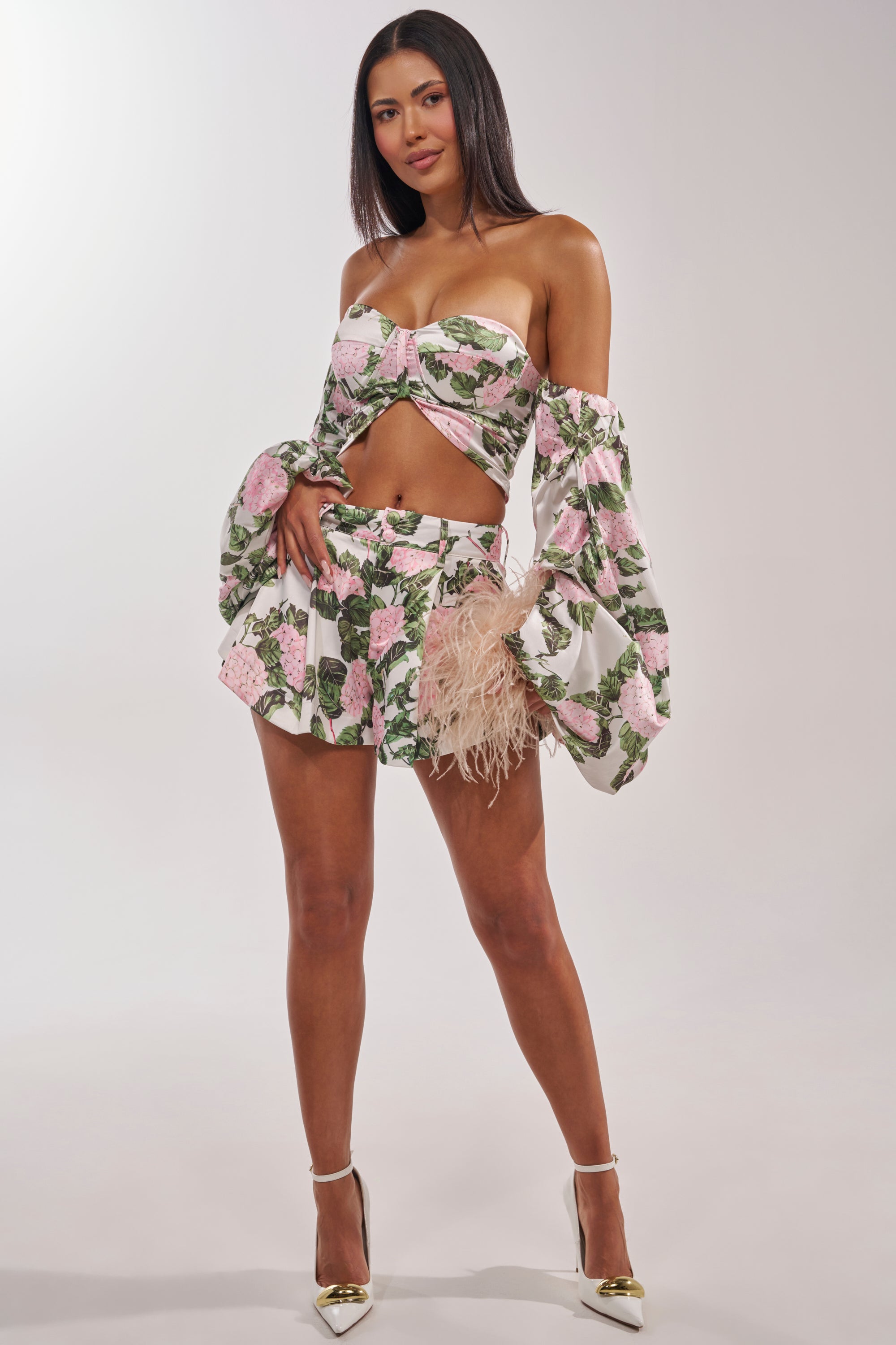 A woman models the ONE OF A KIND FLORAL SHORT with a matching floral off-shoulder crop top, holding a beige feathered clutch and wearing white pointed heels against a plain, light background. 7