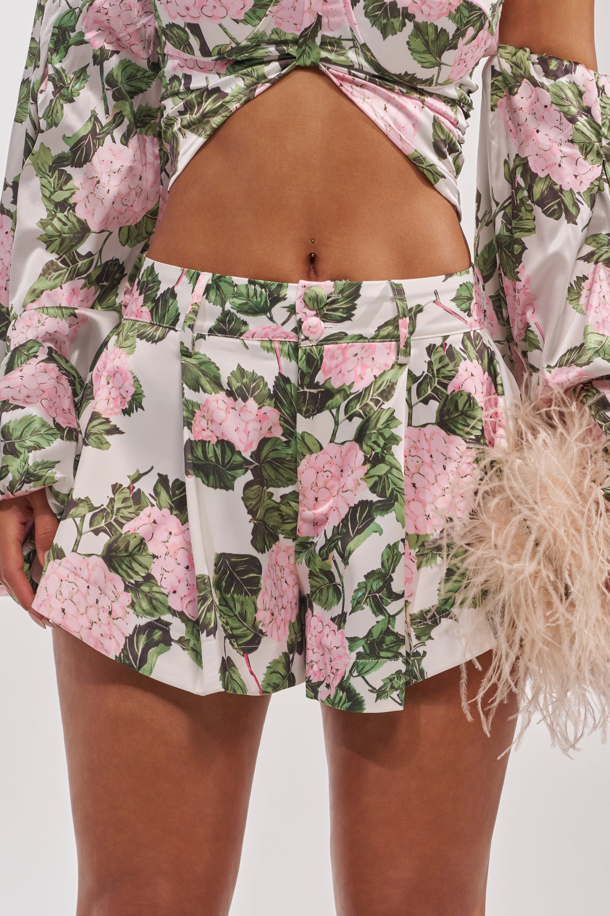 Someone wears the ONE OF A KIND FLORAL SHORT—pleated satin shorts with a pink hydrangea print—paired with a matching crop top featuring a front cutout and long sleeves, accessorized with a beige feathered piece. 8