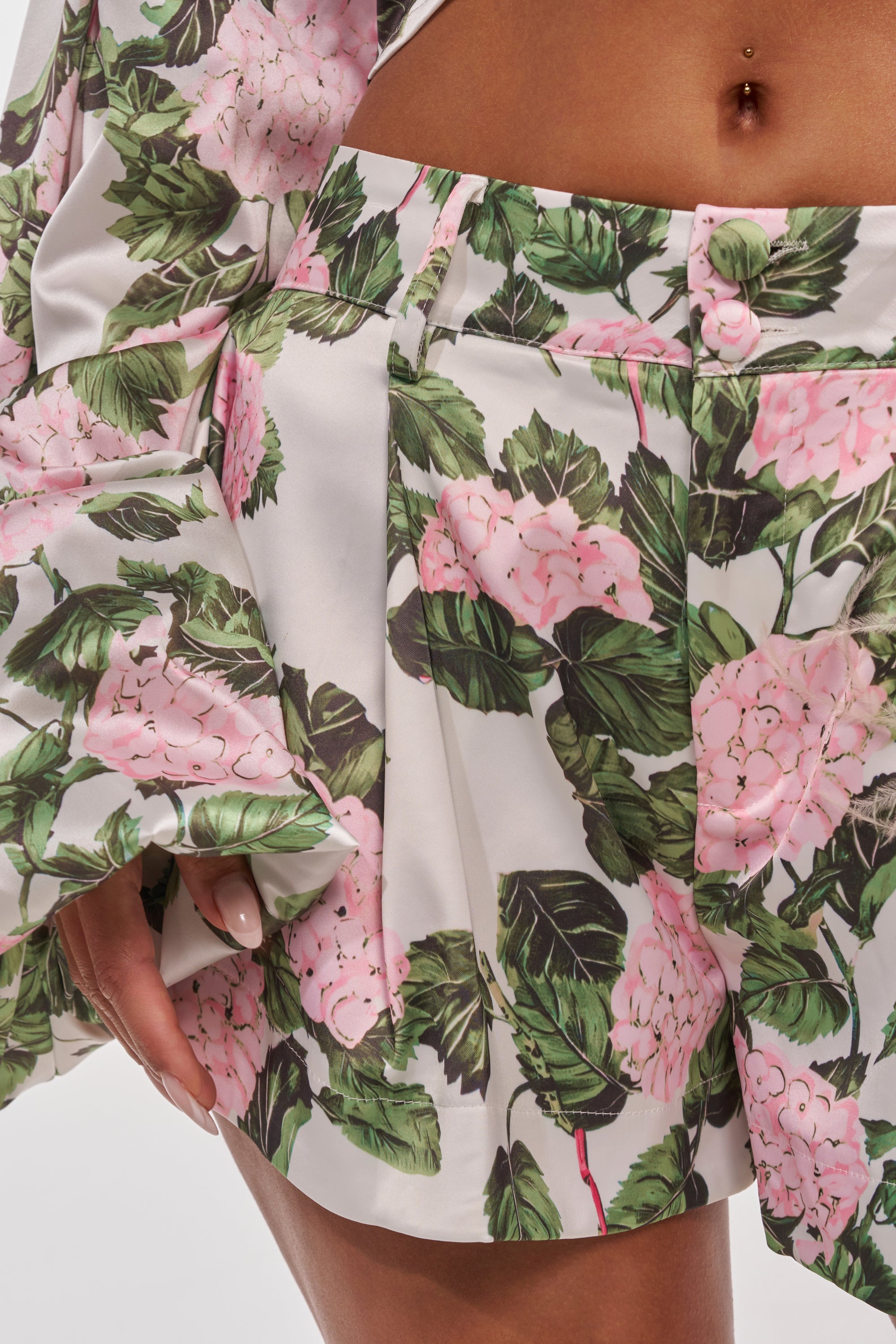 A close-up of a person wearing the ONE OF A KIND FLORAL SHORT featuring pink hydrangea flowers and green leaves on white satin; their hand rests at the side, revealing a midriff with a navel piercing. 9