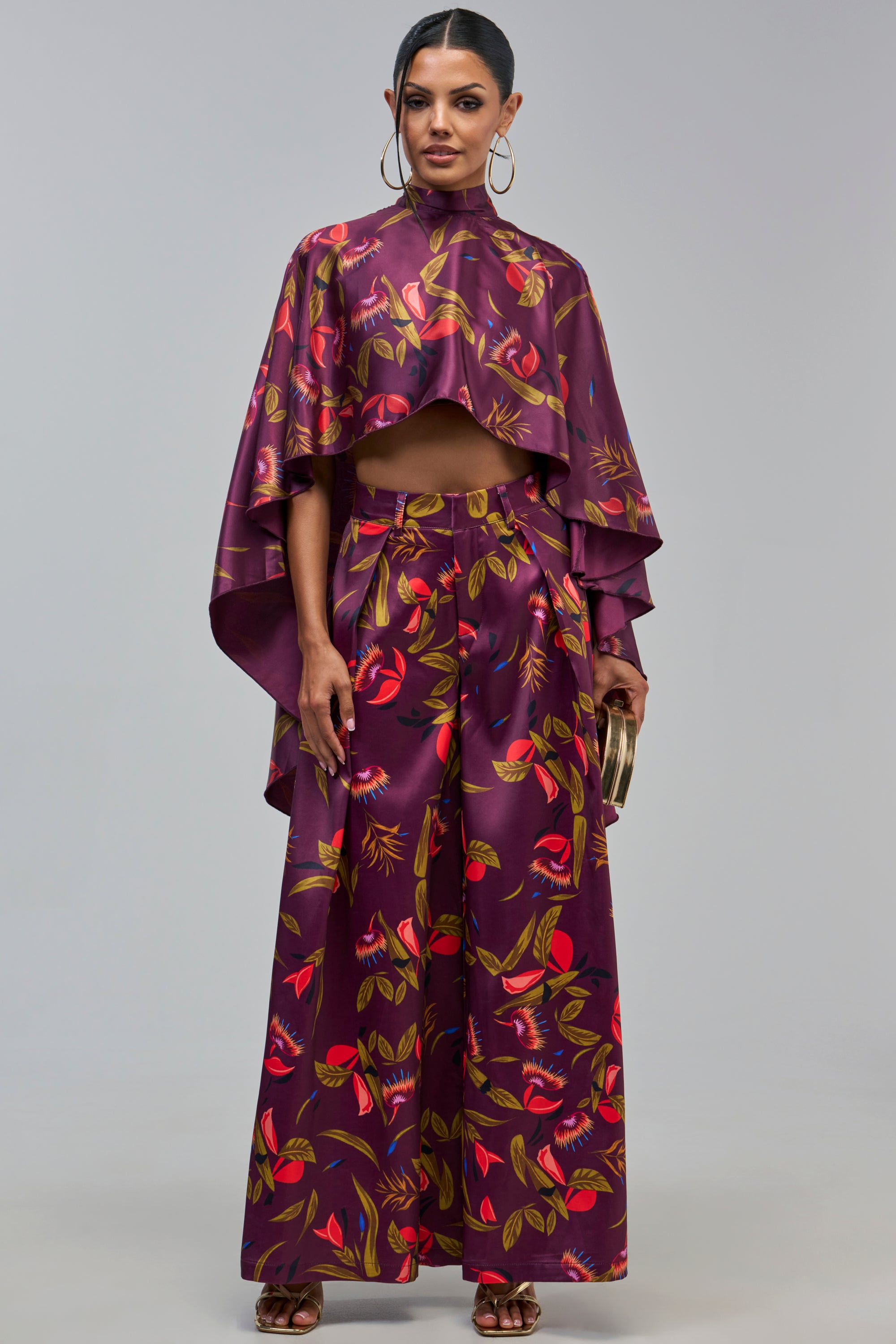 A model wears a matching purple floral print set featuring a cropped mock-neck cape top and the PURPLE RAIN SATIN WIDE LEG PANT.