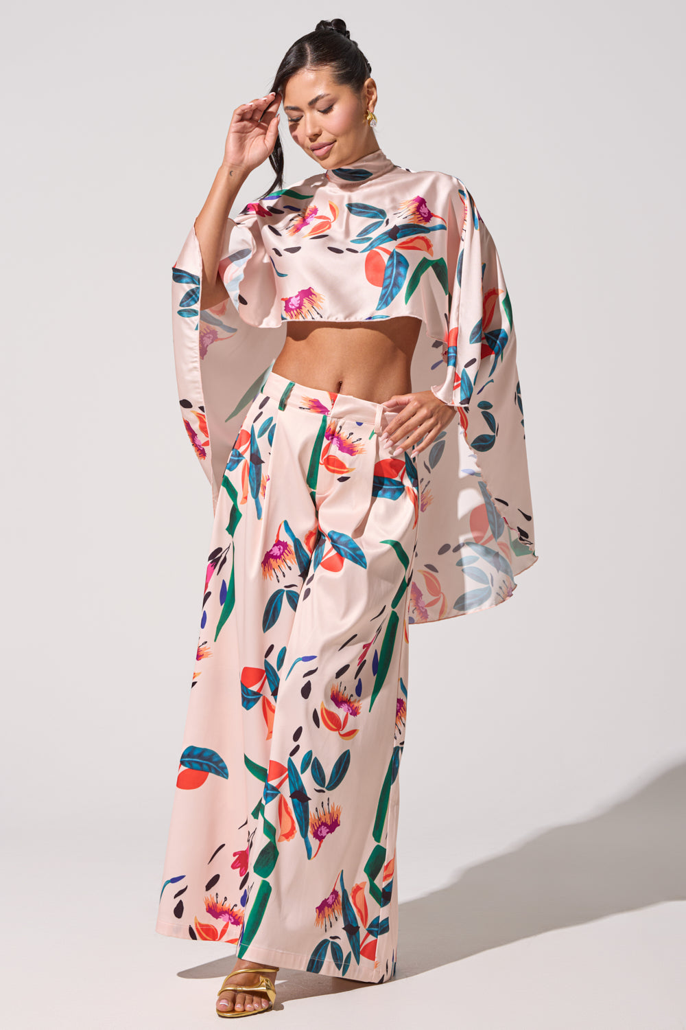 A woman models the CANDY RAIN SATIN WIDE LEG PANT, featuring vibrant multicolored floral and leaf prints. She pairs it with a light pink crop top, gold sandals, gold earrings, and wears her hair partially tied back. 2