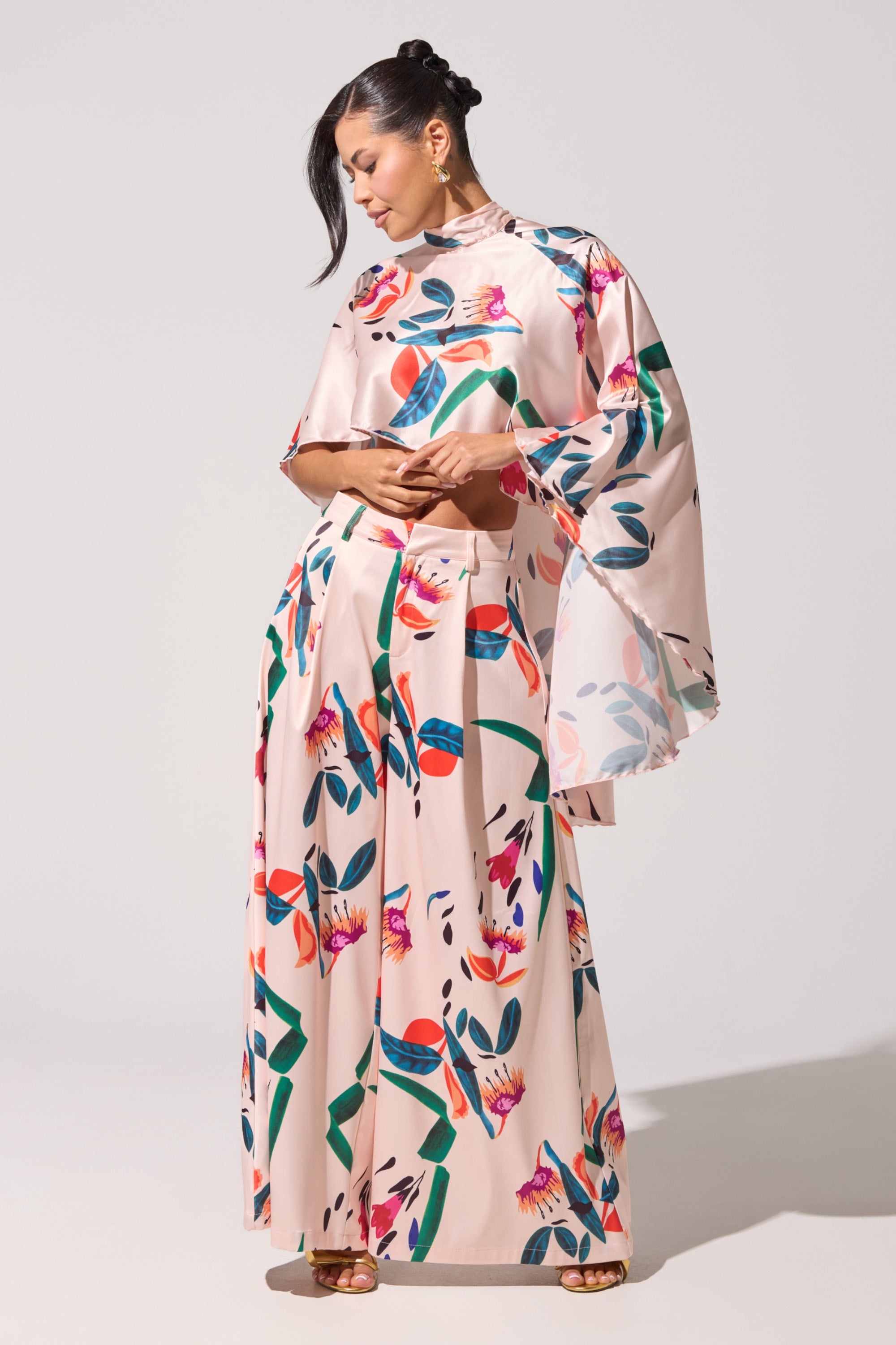 A woman stands against a plain background wearing the CANDY RAIN SATIN WIDE LEG PANT in light pink, featuring bold floral and leaf patterns. She pairs it with a matching wide-sleeved high-neck top, looking down with hands gently clasped. 0