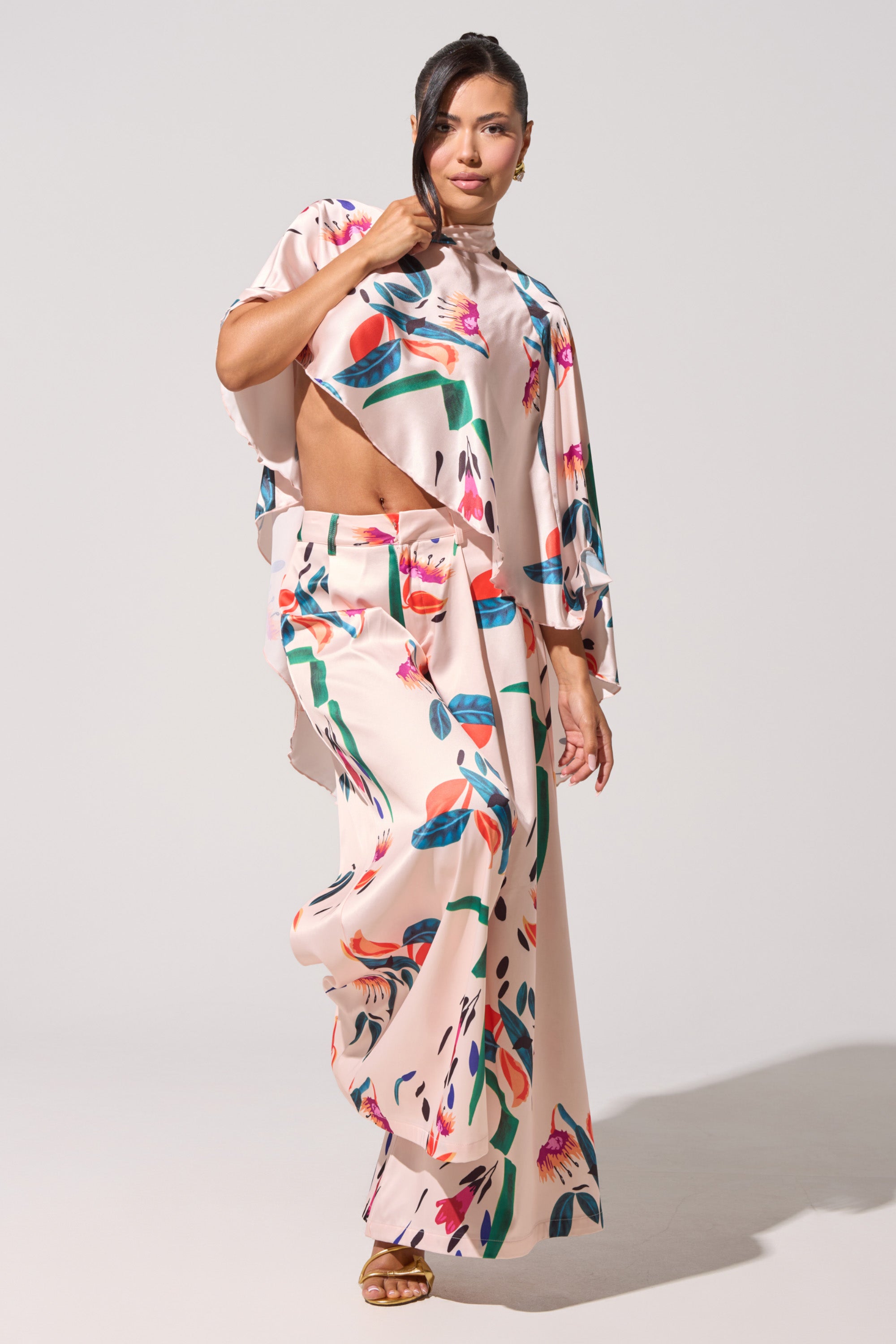 A woman poses confidently in the CANDY RAIN SATIN WIDE LEG PANT, styled with a loose floral top and strappy sandals. Her long hair is pulled back as she stands with one leg forward against a plain background. 1
