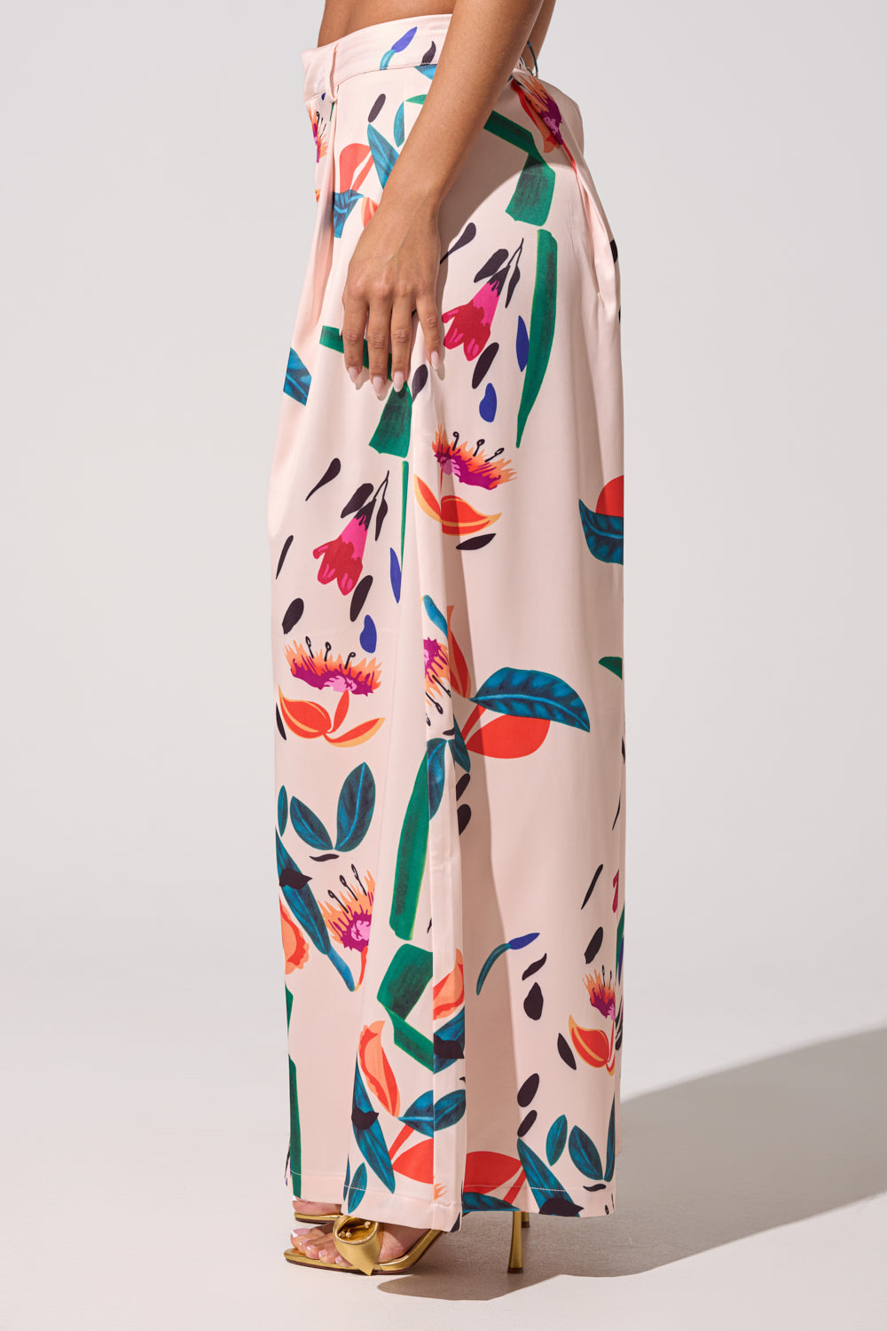 A woman stands in profile on a neutral background, wearing the CANDY RAIN SATIN WIDE LEG PANT—pink floral satin wide-leg pants perfect for spring—paired with gold high-heeled sandals. 4