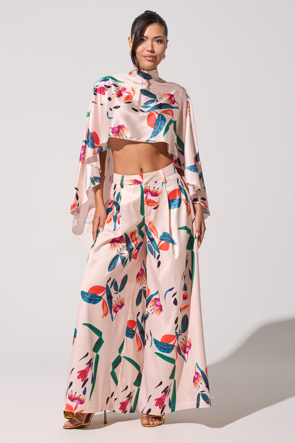 A woman stands confidently against a plain light background, wearing CANDY RAIN SATIN WIDE LEG PANT with a matching pink floral crop top with flowing sleeves and open-toe heels. 6