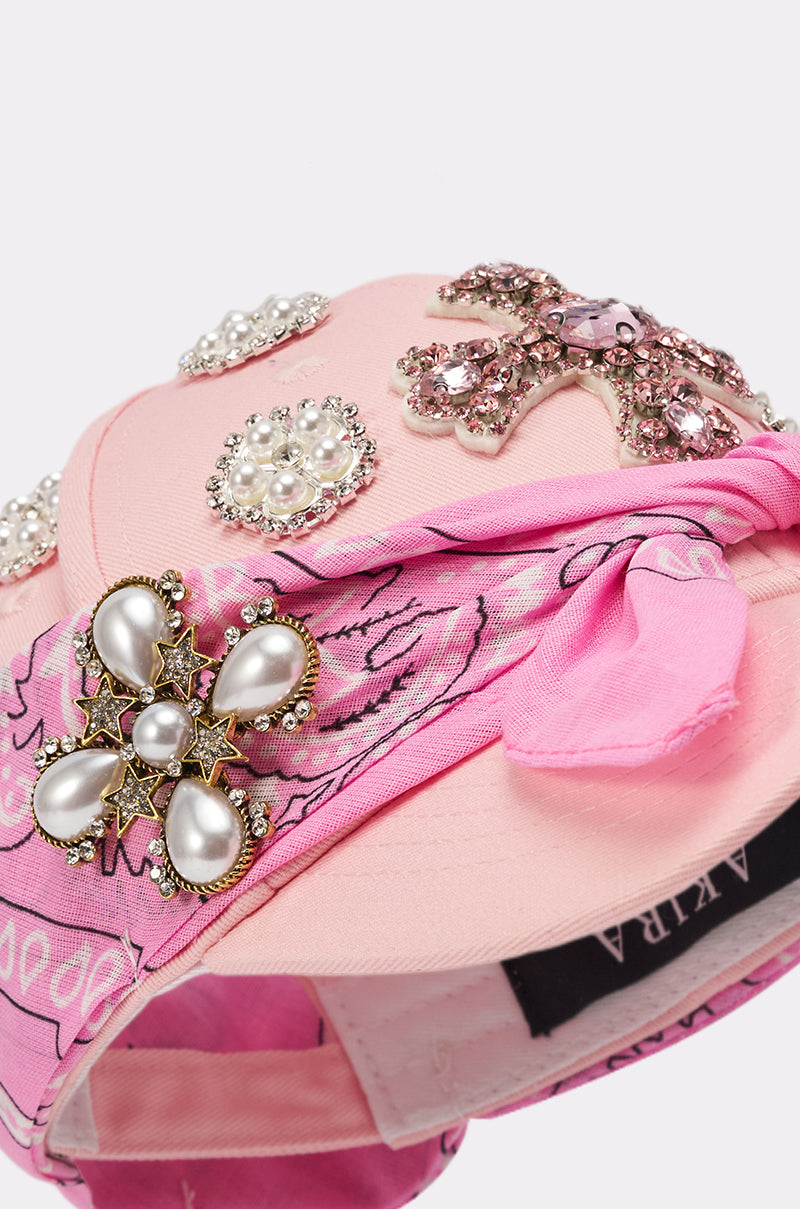 A close-up of the ASTRA PAISLEY BROOCH HAT, a pink rhinestone baseball cap adorned with pearl and rhinestone brooches, a pink bandana around the brim, and a visible black label with white text inside. 4