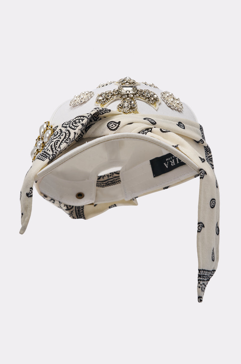 The VIVIENNE PAISLEY BROOCH HAT is a white adjustable baseball cap adorned with rhinestones and brooches, finished with a black and white paisley bandana tied around the brim. 4