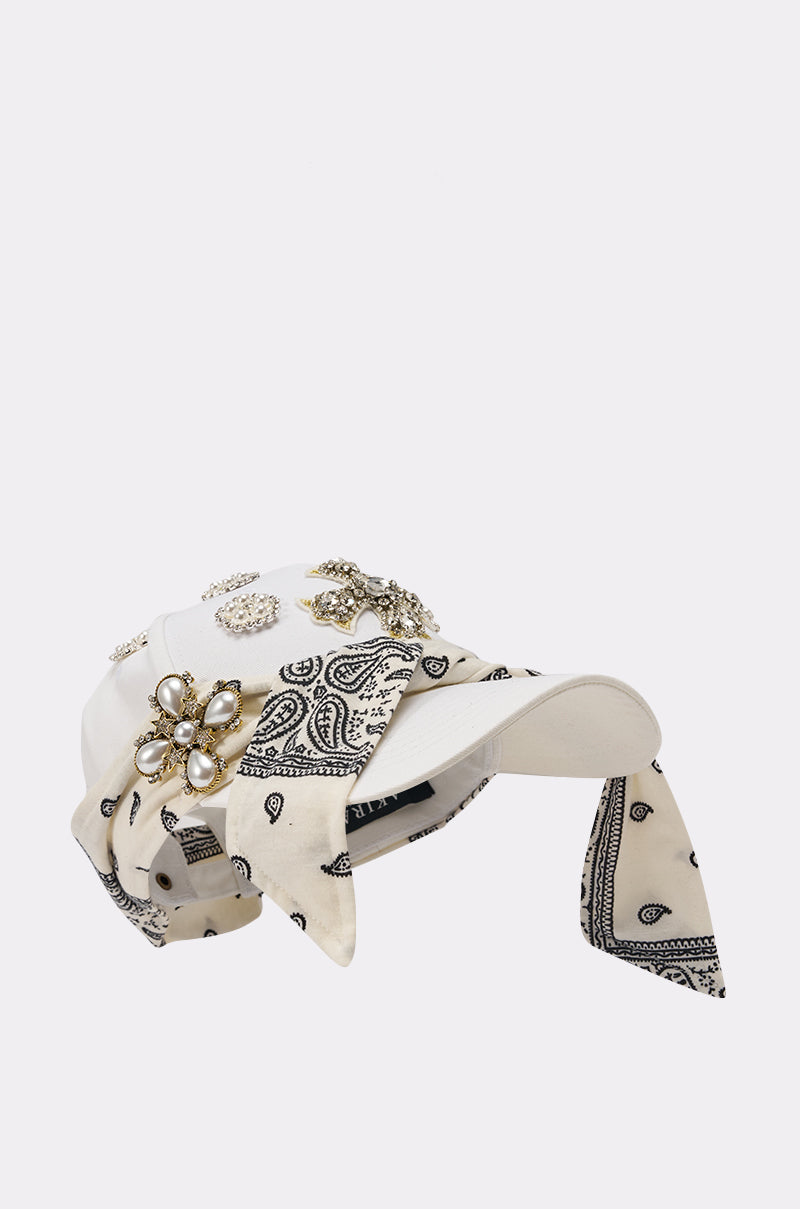 The VIVIENNE PAISLEY BROOCH HAT is a white cap with a black paisley bandana accent, rhinestone pearl brooches on top and sides, and bandana fabric draping elegantly from the back. 5