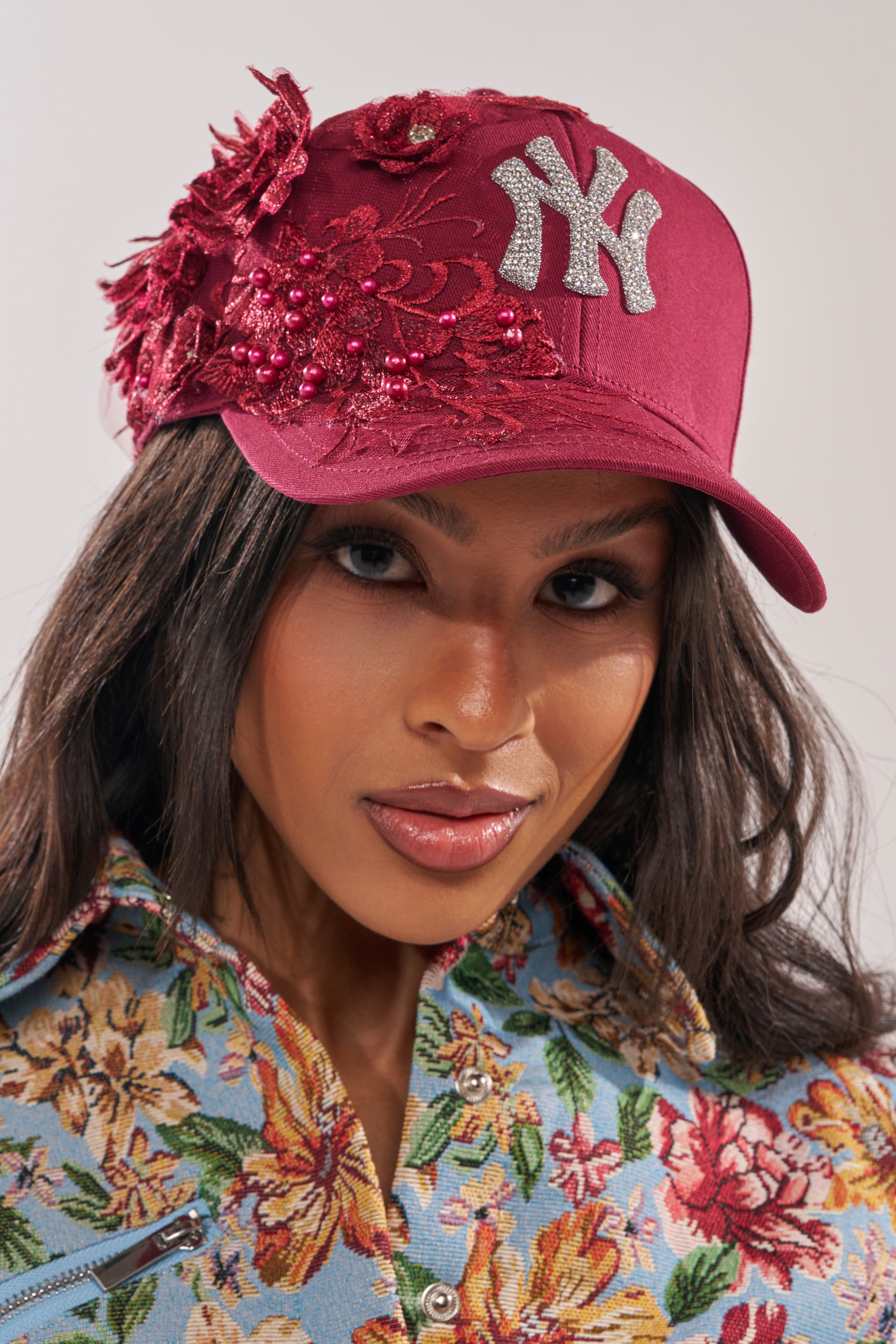 A woman with long dark hair wears the FROM YA CITY APPLIQUE HAT, a maroon New York Yankees cap with floral embroidery and beads, paired with a light blue shirt featuring a colorful floral pattern. 0