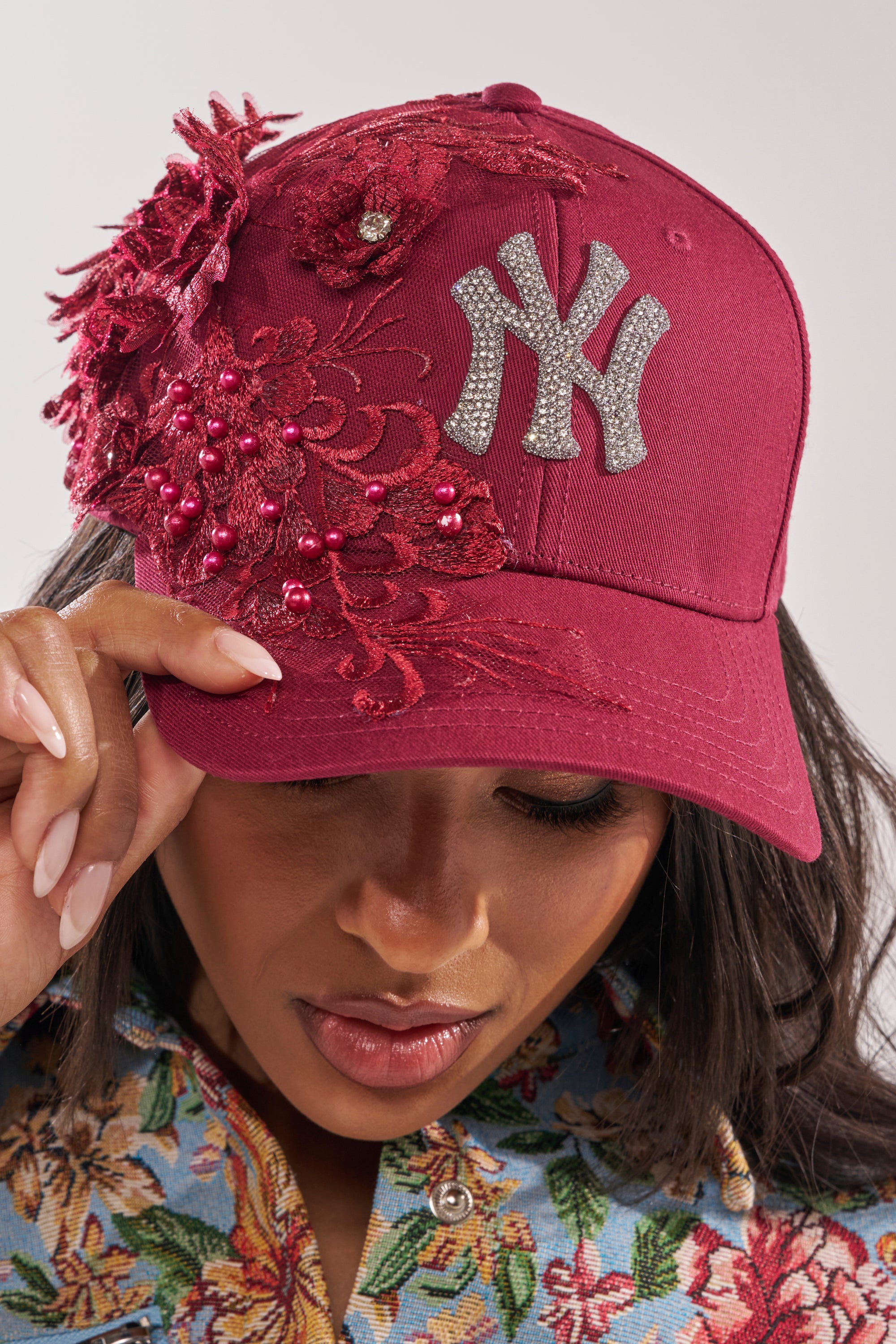 Wearing the FROM YA CITY APPLIQUE HAT, featuring maroon fabric, rhinestones, red lace, and beads, paired with a floral-patterned shirt, this bold accessory stands out as they look down. 1