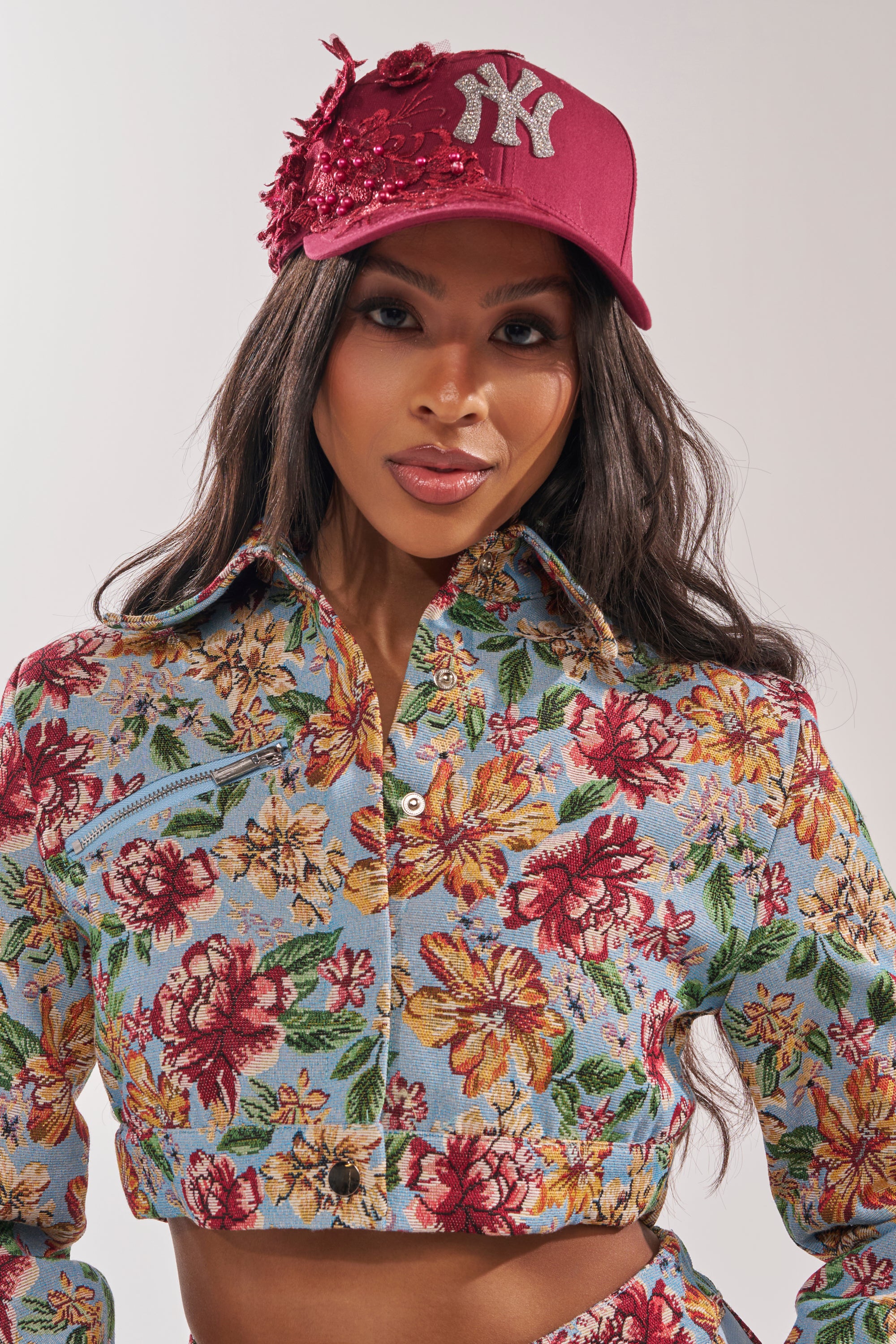 A woman wears the FROM YA CITY APPLIQUE HAT featuring rhinestones and floral details, paired with a cropped, long-sleeve floral top with a blue background, standing against a plain light backdrop to complete her statement look. 6