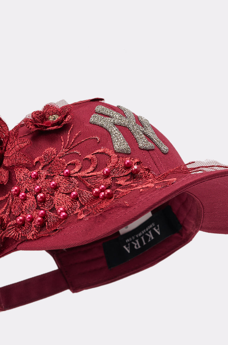 A close-up of the FROM YA CITY APPLIQUE HAT in maroon, featuring decorative red embroidery, beaded accents, and a glittery silver "NY" logo. The inside label reads "AKIRA. 5
