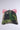 The IM LITERALLY OUTSIDE BOW SNAPBACK, with its vibrant green and brown camo pattern, prominently features a pair of large, bright pink satin bows attached to the back. This creates a striking contrast between the rugged and playful elements. The adjustable strap ensures a perfect fit against a plain, light gray background.