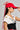 A woman wearing the BIGGER IS BETTER BOW SNAPBACK in red, paired with large hoop earrings and a white top featuring a black design. Her top is adorned with a small badge and the text "2024." She stands sideways, adjusting her bow snapback with her hand.