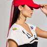 A woman wearing the BIGGER IS BETTER BOW SNAPBACK in red, paired with large hoop earrings and a white top featuring a black design. Her top is adorned with a small badge and the text "2024." She stands sideways, adjusting her bow snapback with her hand.