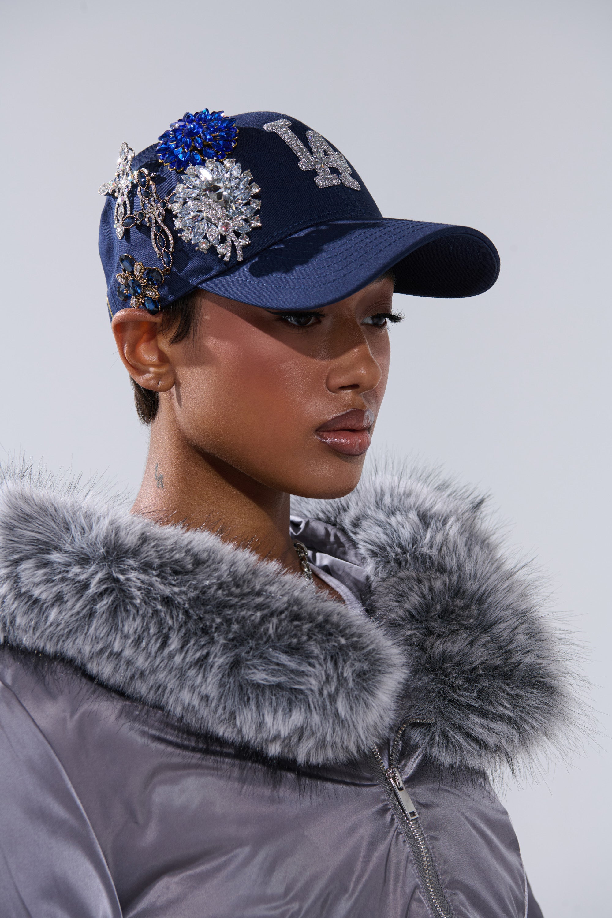 A person in the IN THE NIGHT CHARM HAT, a navy blue baseball cap with sparkling brooches, and a shiny gray jacket with a fluffy fur collar, looks serious against a plain background. 0