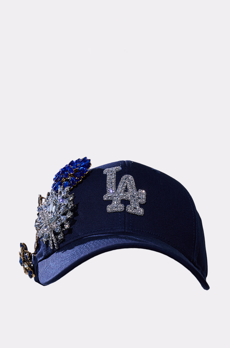 The IN THE NIGHT CHARM HAT is a navy blue baseball cap with a glittery "LA" logo on the front and bold rhinestone embellishments on the left side. 2