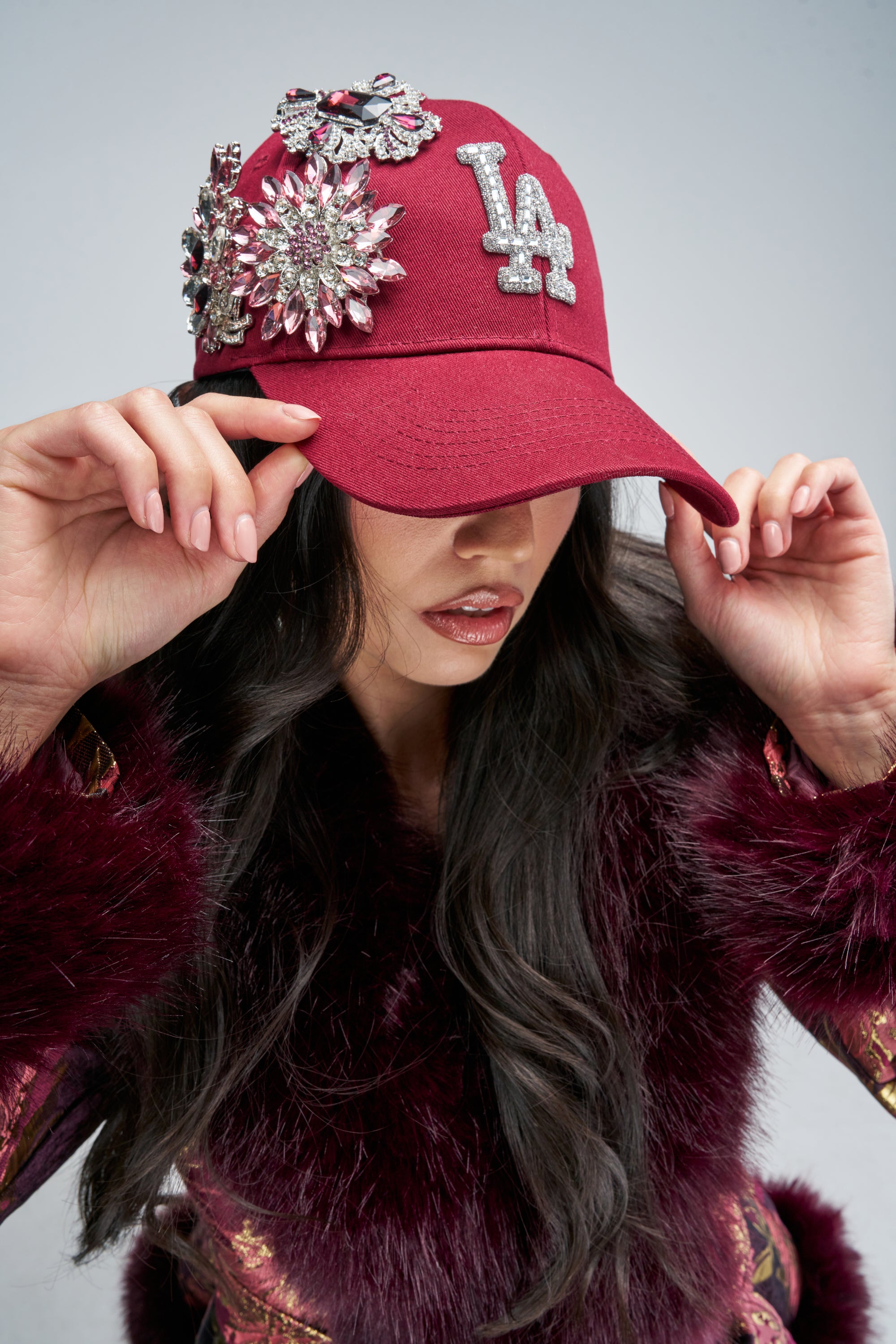A person wearing a BROOCH BABE EMBELLISHED HAT, a burgundy baseball cap featuring a rhinestone “LA” logo and large crystal brooches.