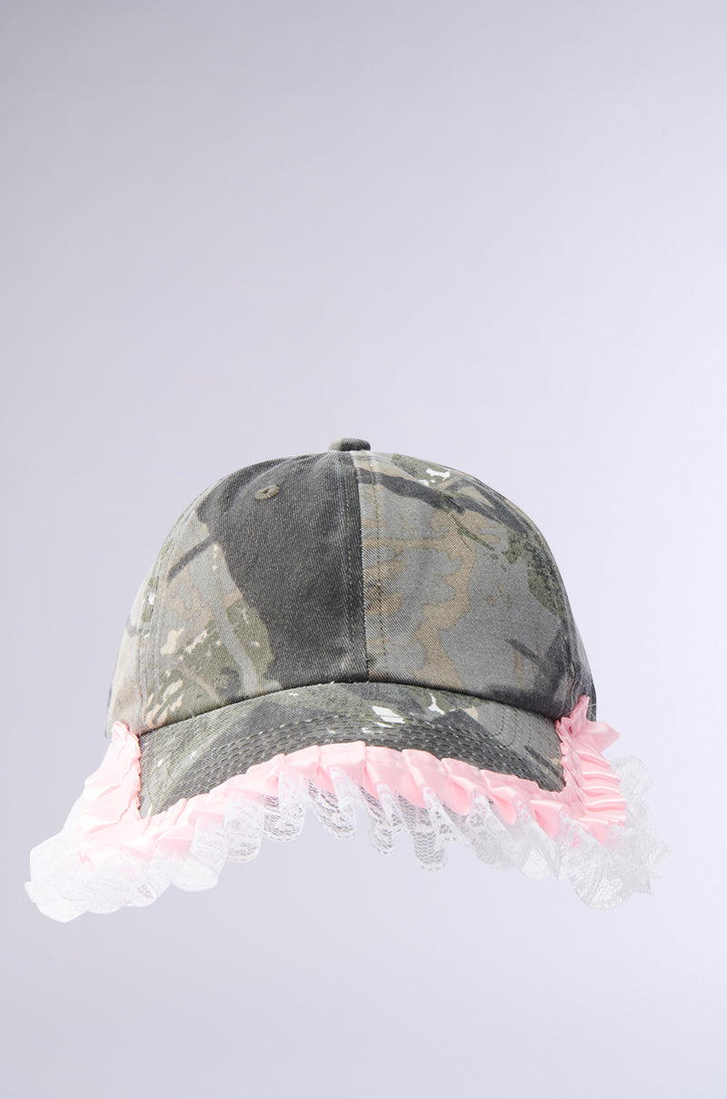 The GIRLY VIBES CAMO HAT showcases a camouflage print with a pink ruffle and white lace trim along the brim, adding feminine flair against a plain light gray background.
