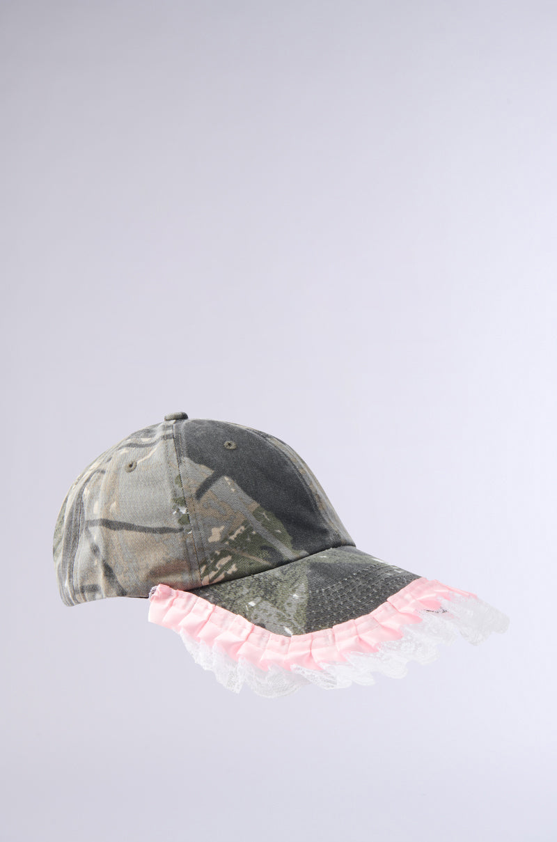 The GIRLY VIBES CAMO HAT features camo print with pink frills and white lace trim on the brim, bringing feminine flair against a plain light gray background.