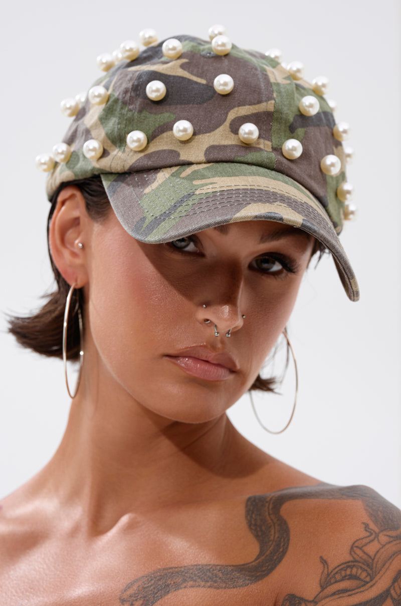 A woman with short dark hair and a neutral expression wears the PEARL PLEASE CAMO HAT, large hoop earrings, and a septum piercing; her visible shoulder tattoo completes the look. 0