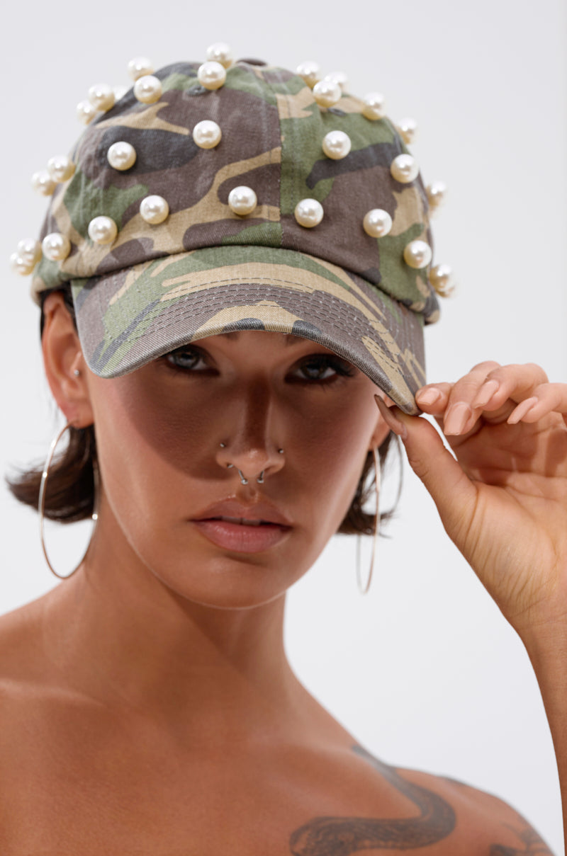 Wearing the PEARL PLEASE CAMO HAT, a person with hoop earrings and multiple nose piercings holds the brim and looks directly at the camera. 1