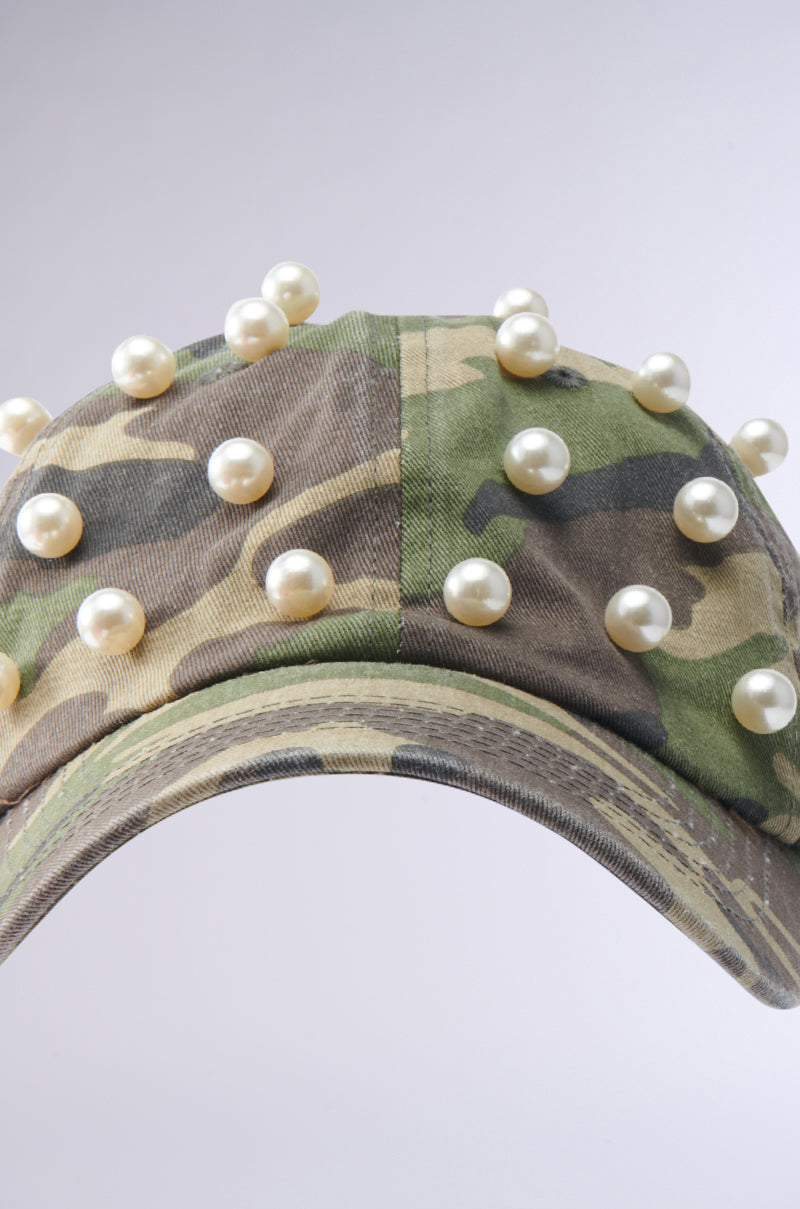 The PEARL PLEASE CAMO HAT showcases a classic camouflage print, highlighted by large white pearls arranged evenly on the crown and brim, all set against a light background. 4