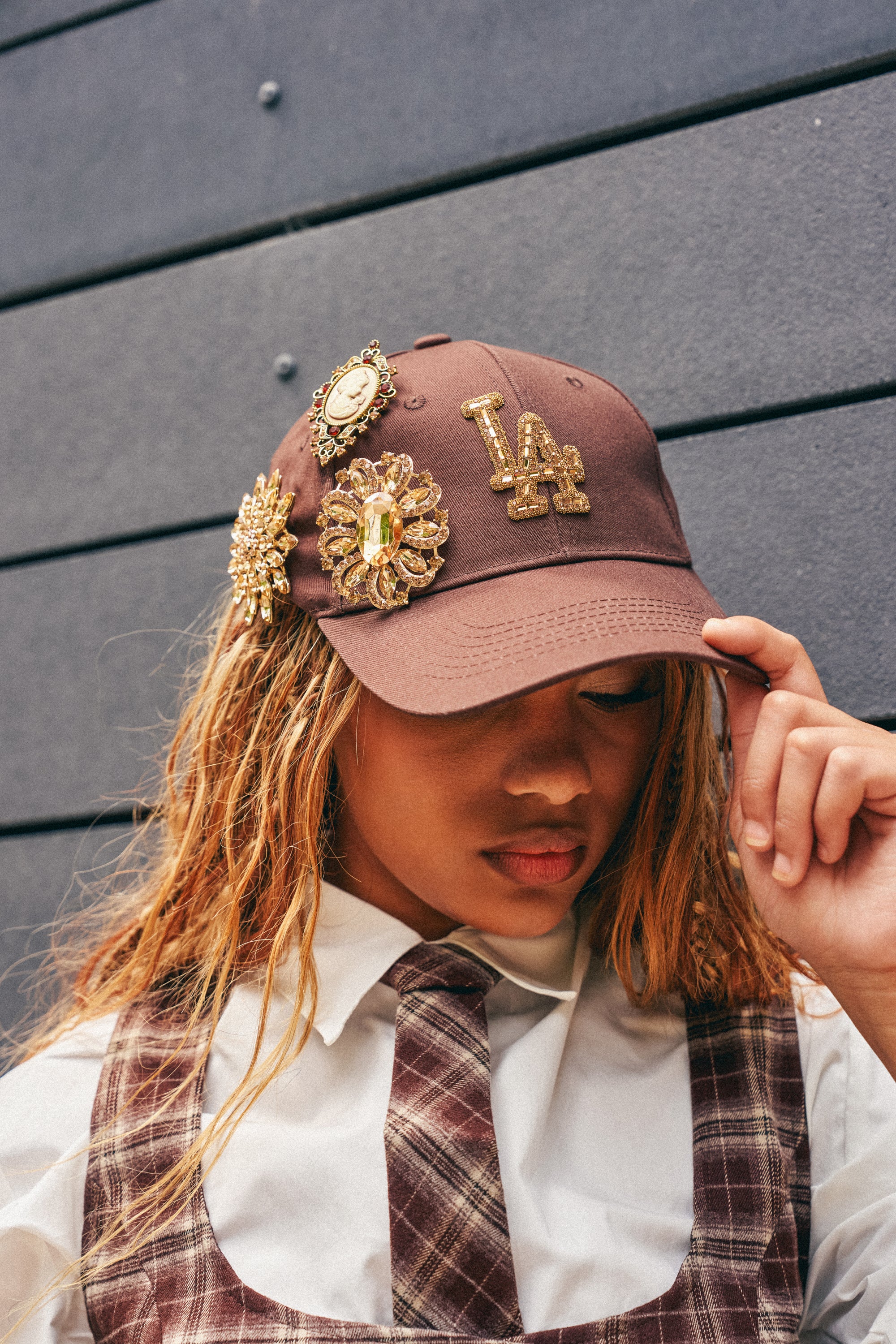 Wearing the LA BADDIE HAT IN BROWN, a person with blond hair partially covering their face stands against a dark wall, looking down, dressed in a plaid shirt adorned with gold brooches.