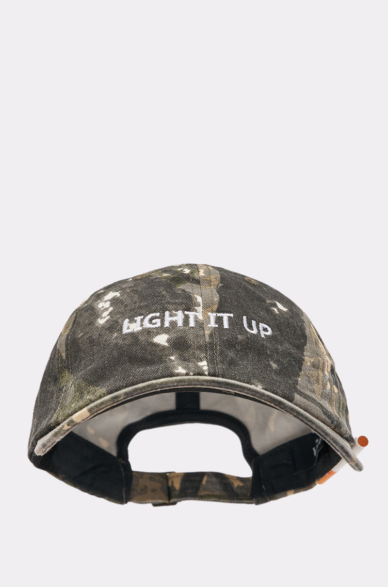 The LIGHT IT UP FAUX CIG HAT, a camo baseball cap with "LIGHT IT UP" embroidered in white on the front, is displayed facing forward against a plain light gray background, highlighting its street style vibe. 4